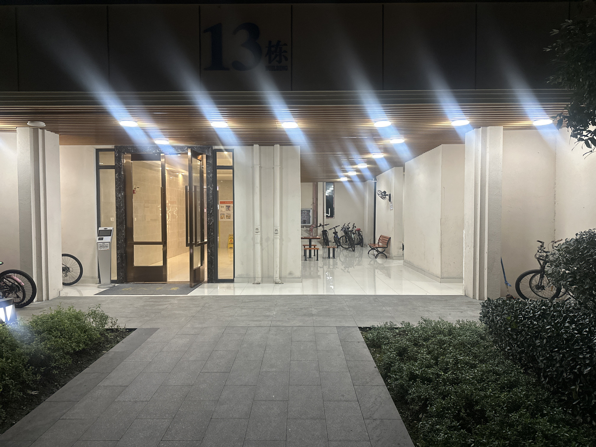 The image displays the illuminated entrance of a modern building at night. The scene is dominated by the building's ground floor facade, featuring a central glass double-door entrance that is slightly open, revealing a brightly lit interior.
Above the entrance, the number "13" is prominently displayed, followed by the Chinese character "栋" (dòng), meaning "building" or "block." Below this, the word "BUILDING" is faintly visible. A series of bright, circular lights are embedded in the slatted ceiling above the entrance, casting strong light rays onto the surrounding area and creating reflections on the clean, tiled floor inside.
To the left of the main doors, a bicycle is parked against a white pillar, and a tall, slender kiosk stands nearby. To the right, the covered entryway extends, providing a sheltered space where multiple bicycles are neatly parked against a wall, alongside a small wooden bench and a table with chairs. A wall-mounted fan is visible further down this corridor.
The foreground features a dark gray tiled pathway leading directly to the entrance, bordered by well-maintained green bushes on both sides. Another bicycle is partially obscured by dense foliage on the far right. The time of day is clearly night, with the artificial lighting starkly contrasting against the dark sky. No individuals are present in the scene, and no specific activity is taking place. The architecture and presence of numerous bicycles suggest a residential or commercial building in an urban environment, consistent with the provided location context of Chongqing High-tech Zone, China.