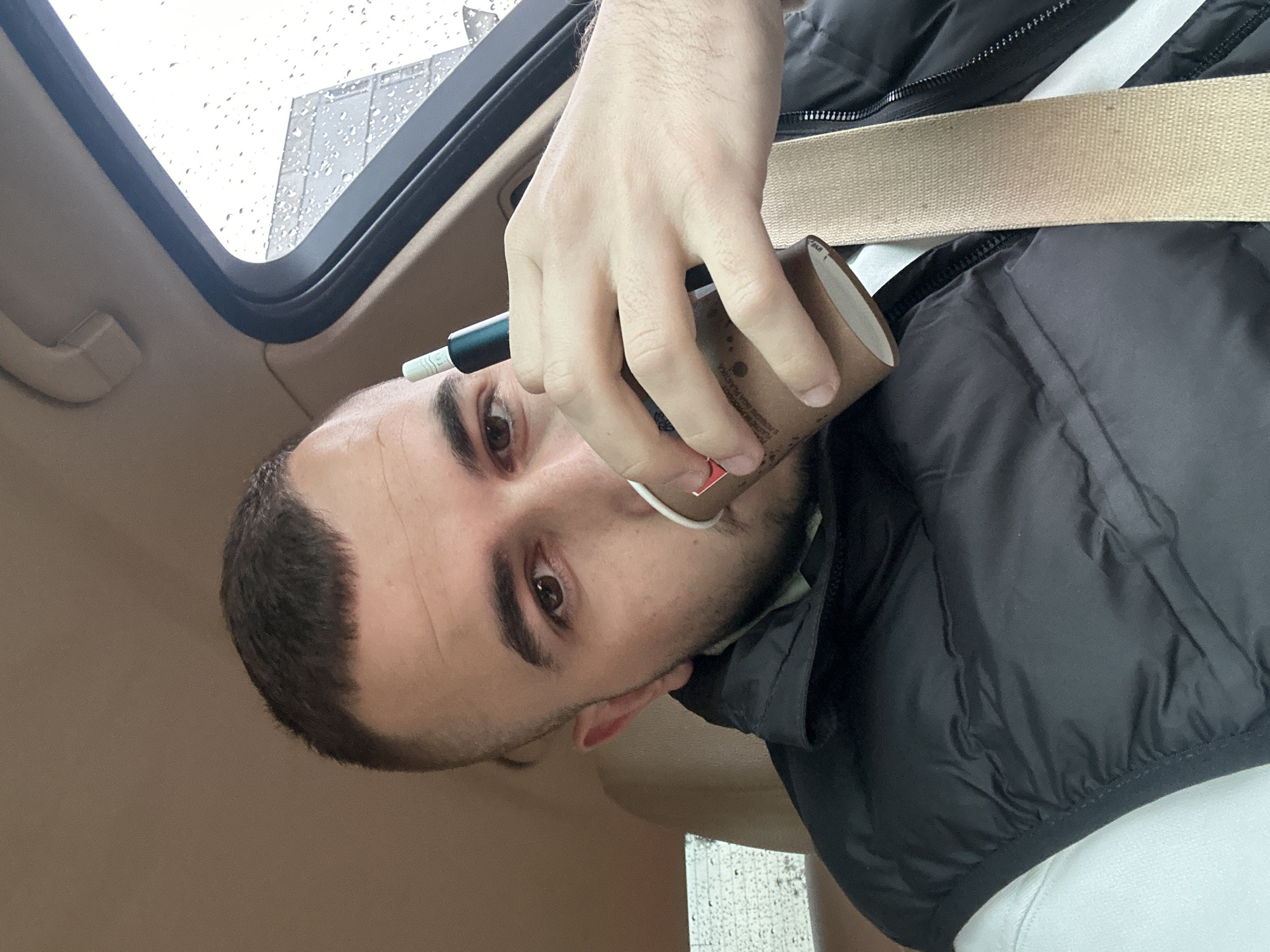 A young man, likely in his 20s or 30s, is captured in a close-up, chest-up shot inside a car, facing the camera. He has short dark hair, a clean-shaven face, and dark eyes. He is wearing a black puffer vest over a light-colored shirt, with a tan seatbelt diagonally across his chest. His right hand holds a small, brown paper cup, featuring a red label and some indistinct small text, positioned near his mouth. He is simultaneously holding a dark, pen-like electronic device, possibly a heated tobacco product, with a white tip, in the same hand, near the cup.
The setting is the interior of a vehicle, characterized by light beige/tan upholstery visible on the car's headliner and A-pillar. Through an open sunroof or window above and behind him, raindrops are visible on the glass, indicating rainy or recently rainy weather outside. A blurred building or structure can be faintly seen beyond the window. The overall lighting suggests it is daytime, possibly overcast. The man's expression is neutral, and he appears to be looking directly at the viewer. This scene is set in Plovdiv, Bulgaria.