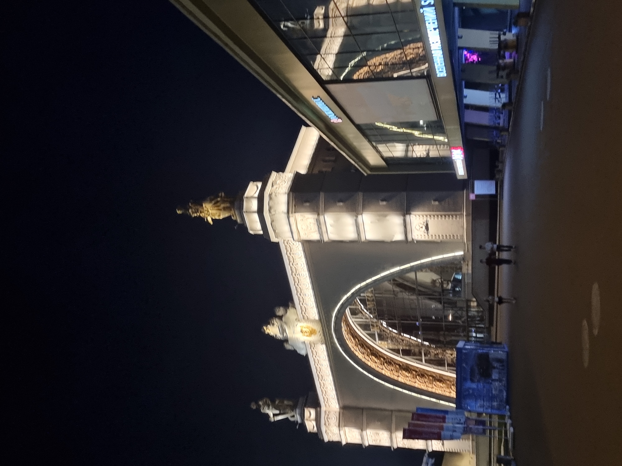 The image captures a nighttime view of an exhibition center in Moscow, Russia, likely the VDNKh (Exhibition of Achievements of National Economy). On the left, a grand archway, illuminated with white lights, frames a large, arched window with ornate golden detailing. Two statues stand atop columns on either side of the archway, and another sculptural element with a golden crest is positioned above the arch. To the right, a modern building with a glass facade reflects the lights and architectural features of the exhibition center. A sign with blue and pink neon letters is visible on this modern structure, along with reflections of lights and possibly other buildings. In the foreground, a paved walkway is visible, with a few people walking in the distance and some blue illuminated structures on the left side. The overall scene is well-lit by artificial lights against a dark night sky.