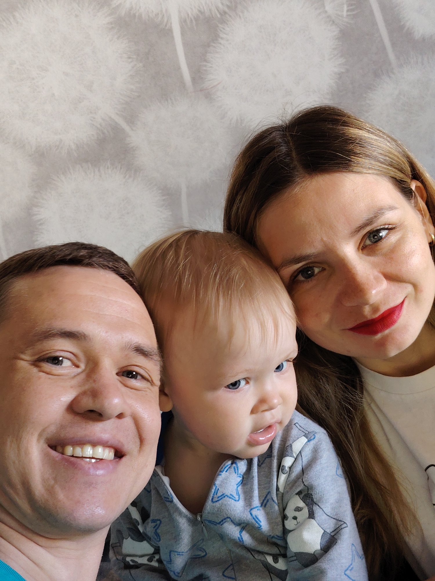 A close-up selfie captures a family of three: a man, a woman, and a baby, positioned closely together in a warm, intimate moment. The man is on the left, smiling broadly with white teeth visible. He has short dark hair and light skin, and wears a blue top. To his right, nestled between him and the woman, is a baby, appearing to be an infant or young toddler. The baby has light blonde hair and striking blue eyes, looking slightly to the right with a neutral to curious expression. The baby wears grey pajamas adorned with blue star outlines and black and white panda patterns. On the far right is the woman, leaning her head gently against the baby. She has long, light brown hair with subtle highlights and wears bright red lipstick, contrasting with her light skin. Her eyes are a light color, and she offers a soft, content smile. A small, gold-colored earring is visible in her right ear. She wears a simple white top. The background is a soft light grey wall, featuring a subtle, repeating pattern of white, fluffy dandelion-like shapes. The lighting is even and natural, suggesting an indoor daytime setting. There is no visible text in the image. The overall impression is one of happiness and familial affection.