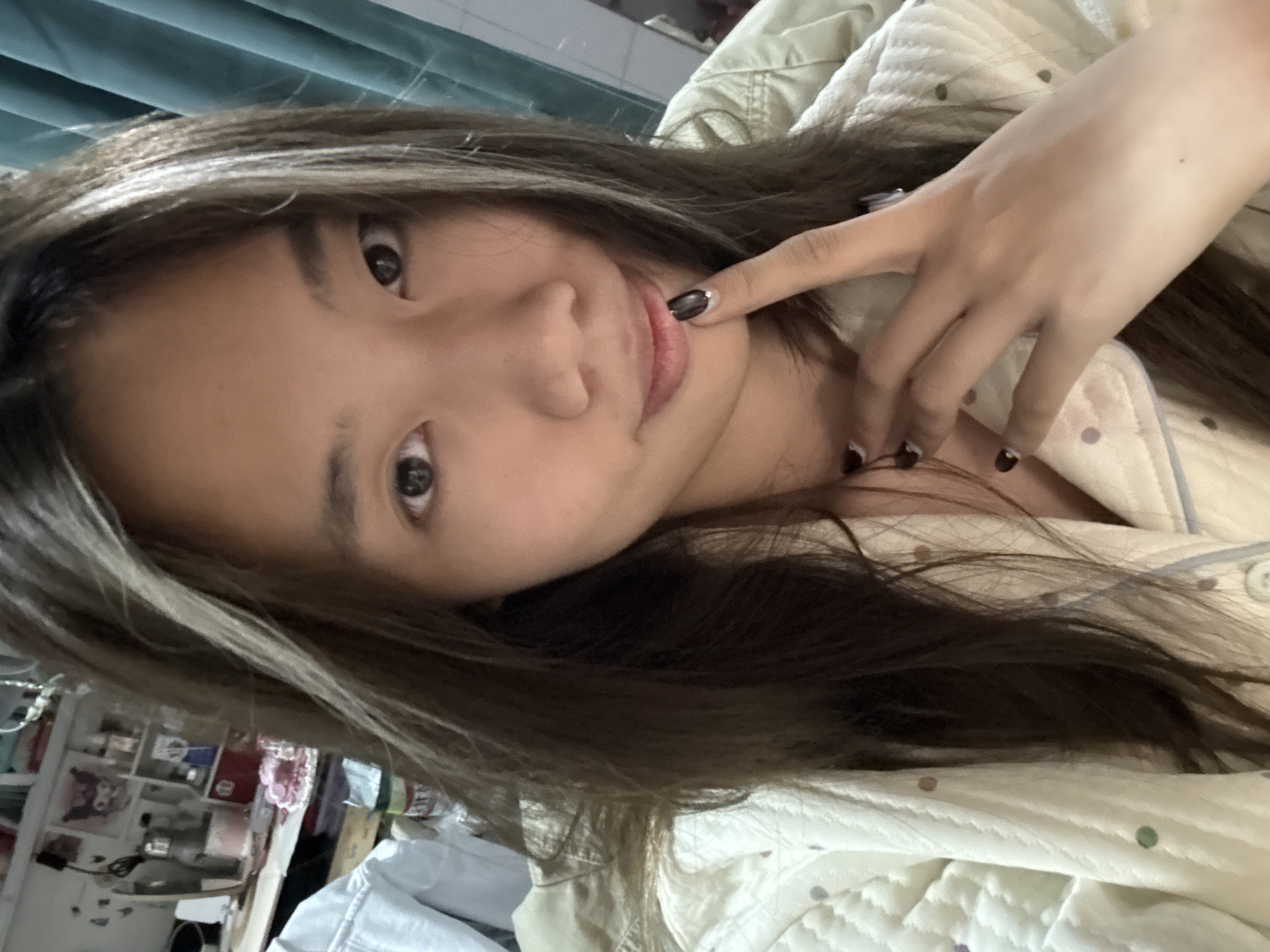 A selfie of a young woman with brown hair and blonde highlights is shown in a close-up, tilted view. She is wearing a pajama top with polka dots. Her left hand is raised to her face, with her index finger near her mouth. Her nails are painted black with a silver accent. In the background, a cluttered shelf is visible, as well as a blue curtain. The photo appears to be taken indoors. There is no information to suggest that this image was taken in the city of Nan'an District, China.
