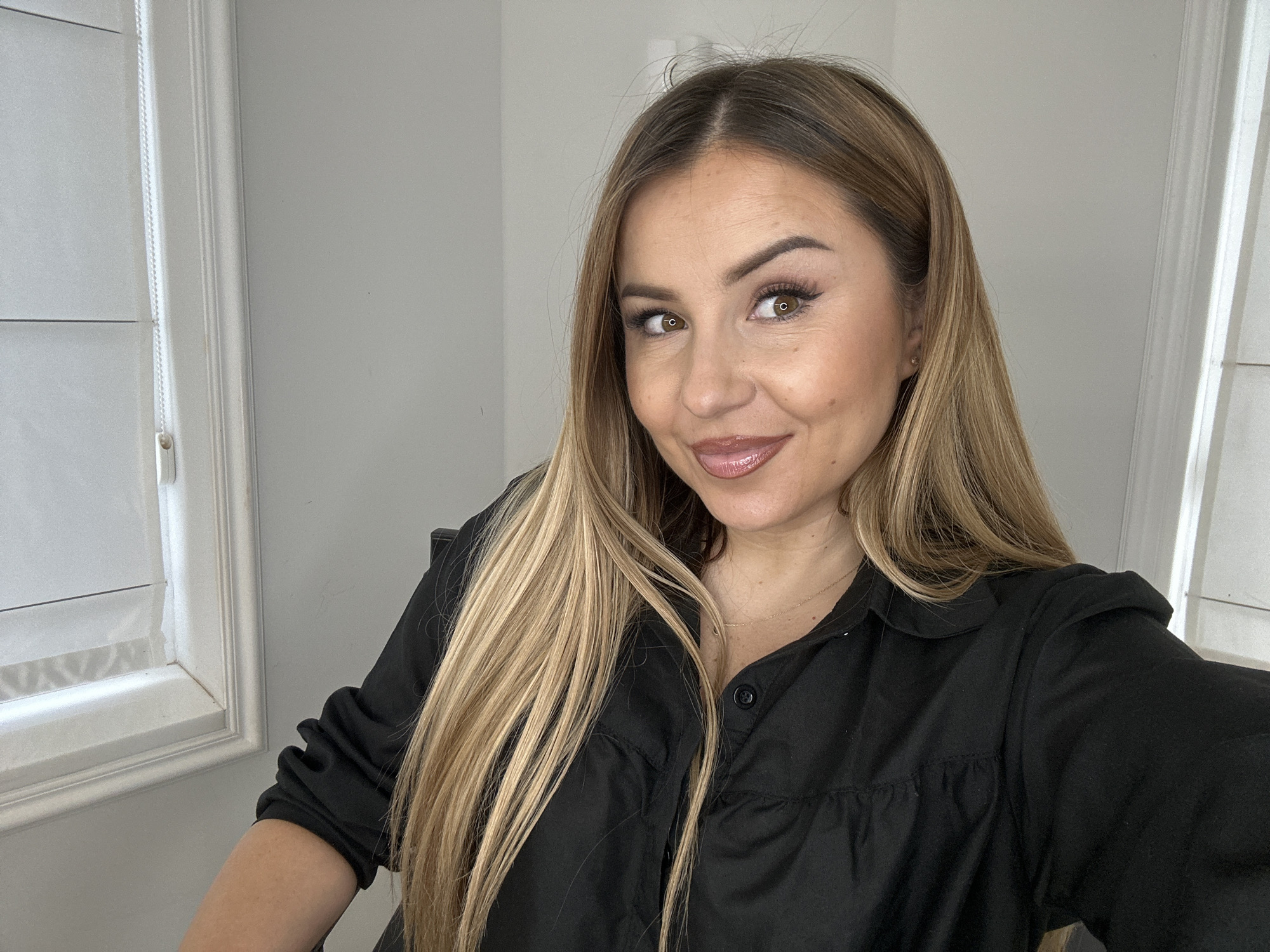 A selfie taken from a slightly high angle shows a woman with long blonde hair and fair skin. She is wearing a black collared shirt. Her makeup includes visible eyeliner, mascara, and lip gloss. She is smiling, with her head tilted slightly to her right. The background is a plain, light-colored wall, with a white-trimmed window and blinds visible to the left of the woman. The scene suggests an indoor setting, possibly a home or office. No specific location in Asenovgrad, Bulgaria is identifiable from the image.