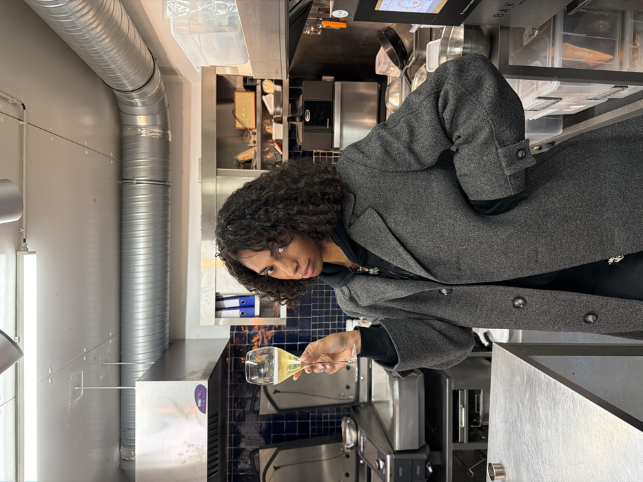 A dark-skinned woman with shoulder-length curly hair, wearing a grey herringbone overcoat over a black hoodie and multiple necklaces, stands facing the viewer. She holds a stemmed glass containing a light yellow, effervescent liquid in her raised right hand, while her left hand rests on her hip. Her gaze is directed forward with a neutral expression.
The scene is set in a commercial kitchen. To the left, stainless steel professional kitchen equipment, including two deep fryers or griddle stations, are visible beneath industrial exhaust hoods. The wall behind these units is clad in dark blue square tiles. To the right of the woman, stainless steel shelving units are visible, holding blue binders, a light brown Marshall brand speaker, and various clear plastic and metal containers. Below the shelving, a dark kitchen appliance and more stainless steel counter space are present. A digital display screen on the far right shows "BAKE" in its lower corner. The ceiling is white, with exposed silver-colored corrugated ventilation ducts running horizontally across the upper frame and fluorescent strip lighting. The specific location within Paris, France, cannot be identified from the visual information provided.