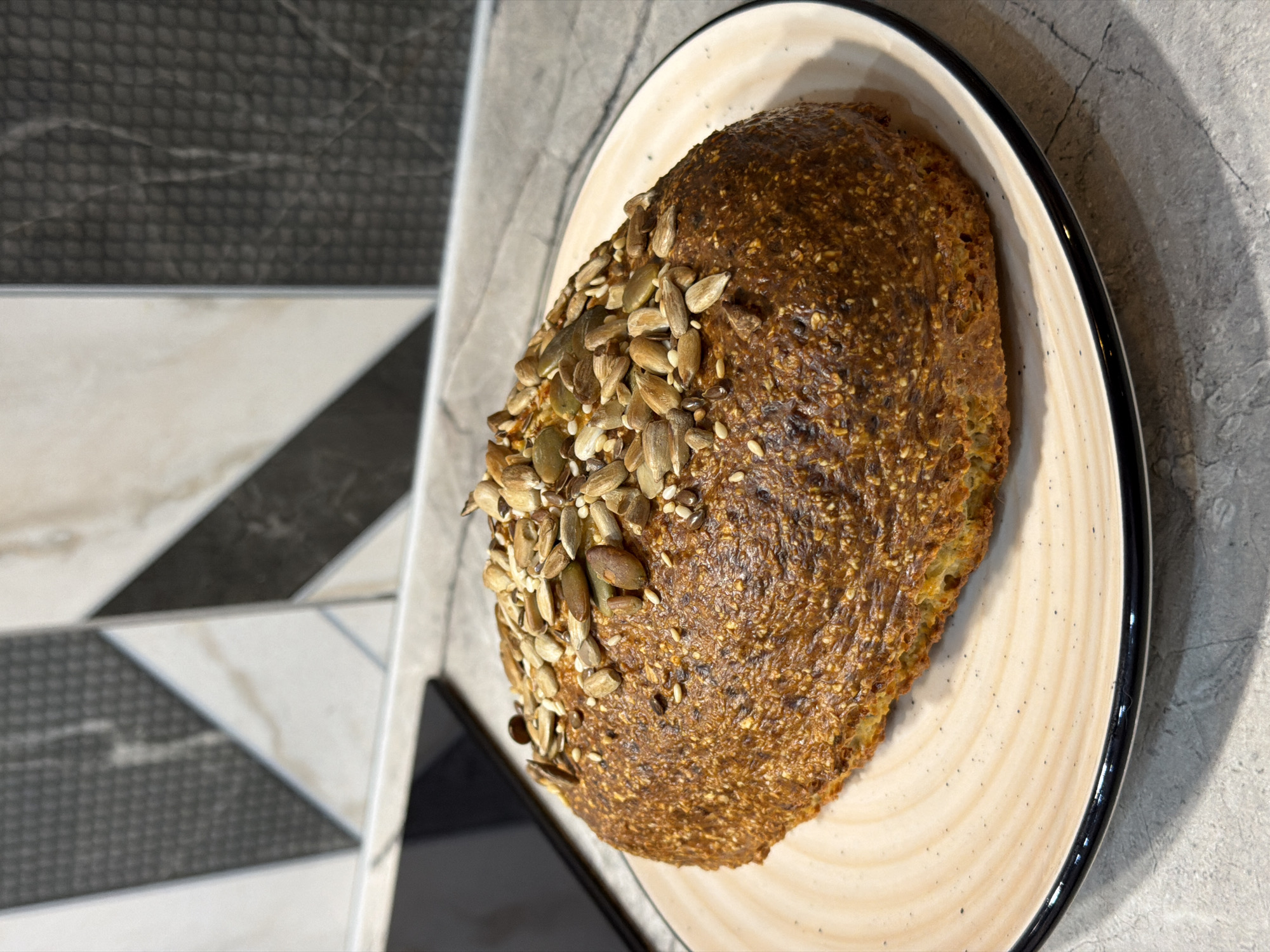 A loaf of brown bread sits on a round plate with a cream-colored base and a black rim. The bread is topped with sunflower and sesame seeds clustered on one side. The plate rests on a gray-patterned surface, with a partial view of a dark object (possibly a phone or tablet) and a geometric tiled surface in the background.
There is no indication of this being taken in Ryazan, Russia.