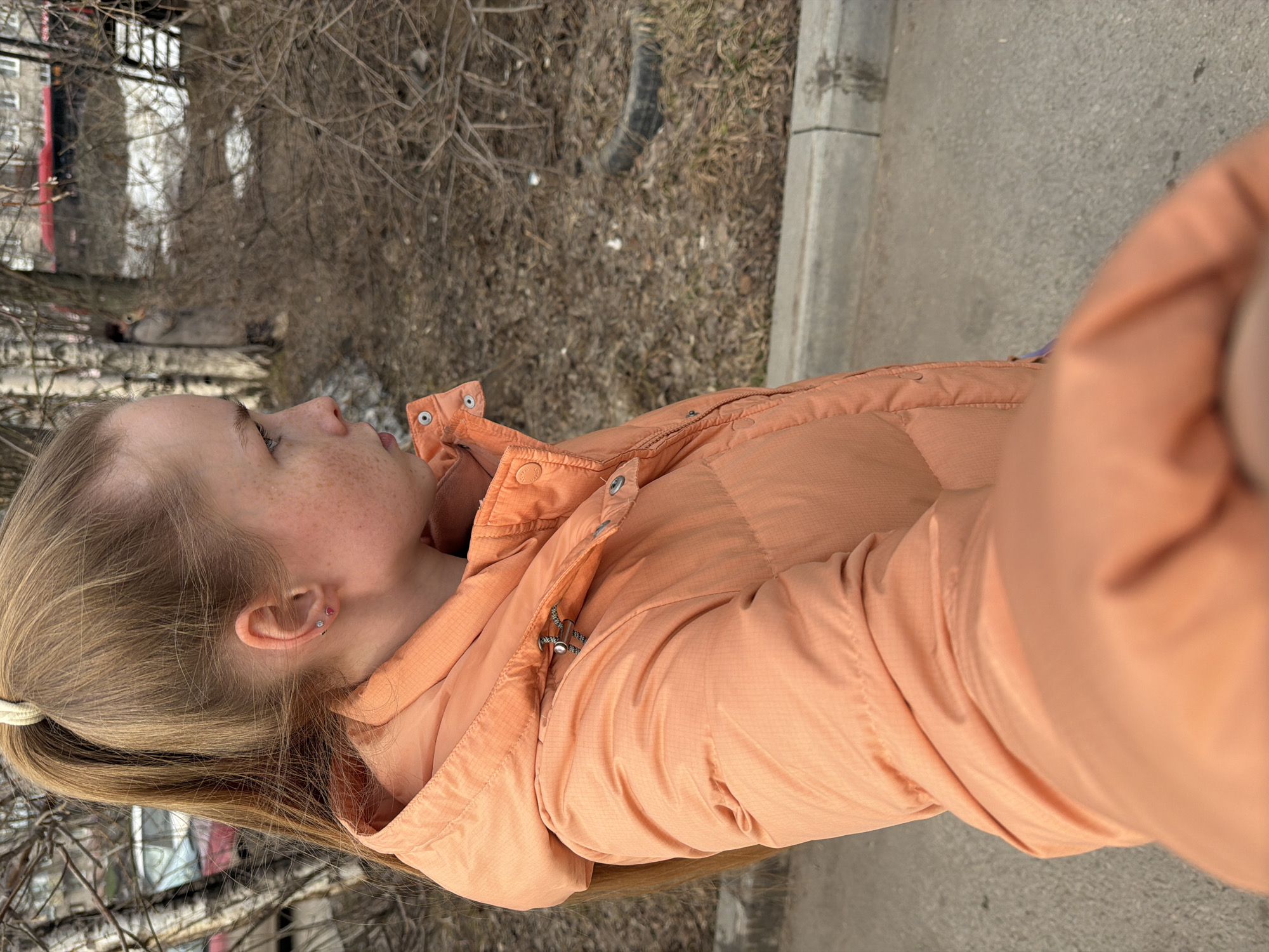 A young, light-skinned person with blonde hair pulled back in a ponytail is standing outdoors, wearing an orange puffy jacket. The individual is facing left in the frame with their head tilted slightly upwards. Their face is visible in profile, showing freckles. The background consists of dry vegetation and trees without leaves. A curb and a paved surface are also visible. Based on available details and the likely location of Berezniki, Russia, the image appears to have been taken near a park or green space in the city.