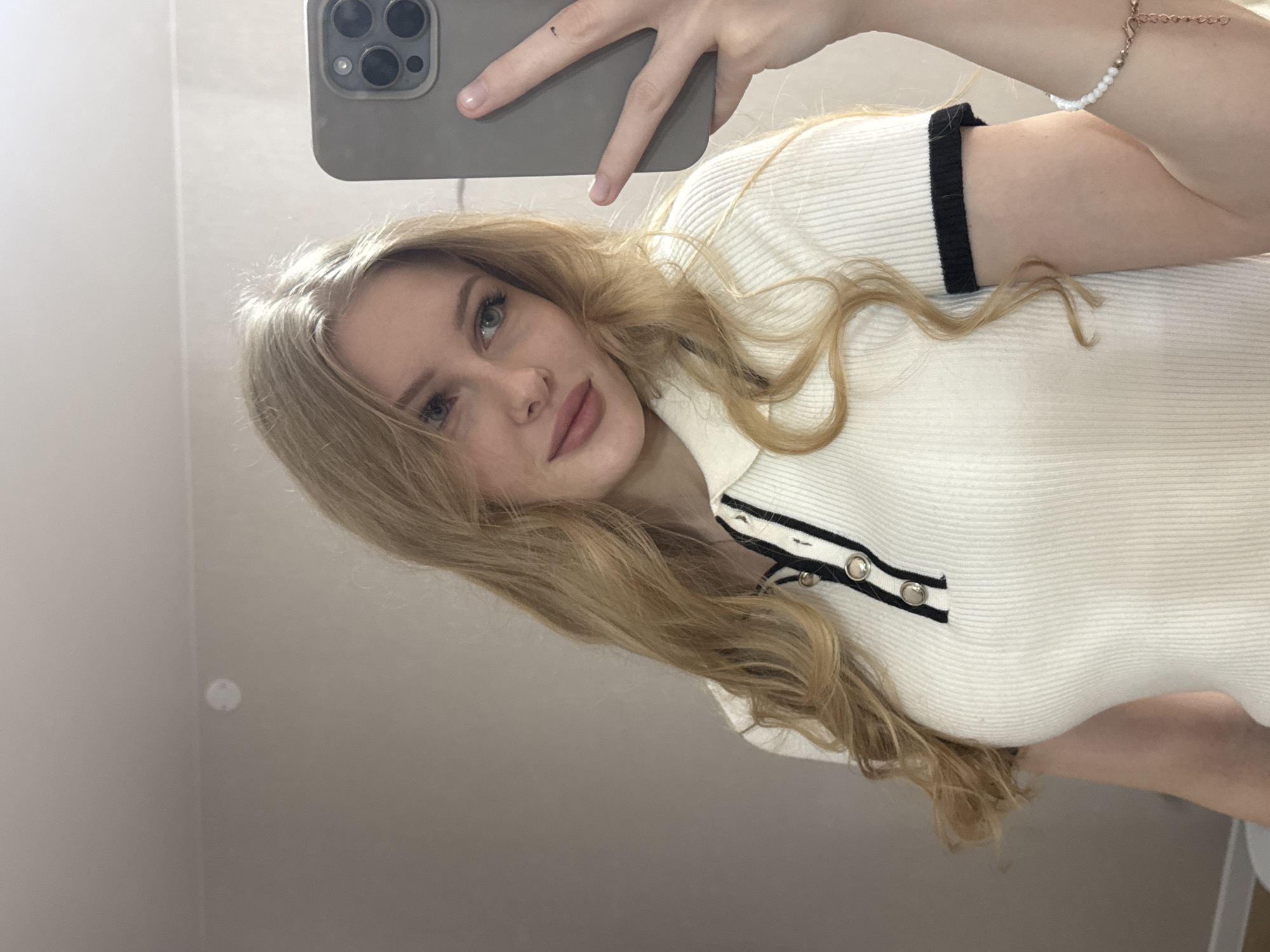 A woman is taking a selfie indoors. She has blonde hair and is wearing a white, ribbed top with black trim. She is holding a grey smartphone up to take the picture. A bracelet is visible on her wrist. The location is a room with plain walls. The photograph does not provide enough details to establish that this photo was taken in Perm, Russia.