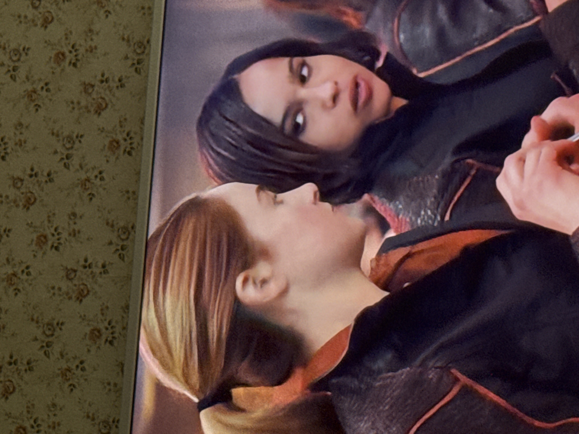 The image appears to be a photograph of a television screen displaying two women. The woman in the foreground has blonde hair pulled back in a ponytail and is wearing a dark colored jacket with orange trim. The woman in the background has short, dark hair and is also wearing a dark colored jacket. The background is slightly out of focus. To the left of the television screen is a wall covered in floral wallpaper. There is no indication in the image that the location is Mytishchi, Russia.