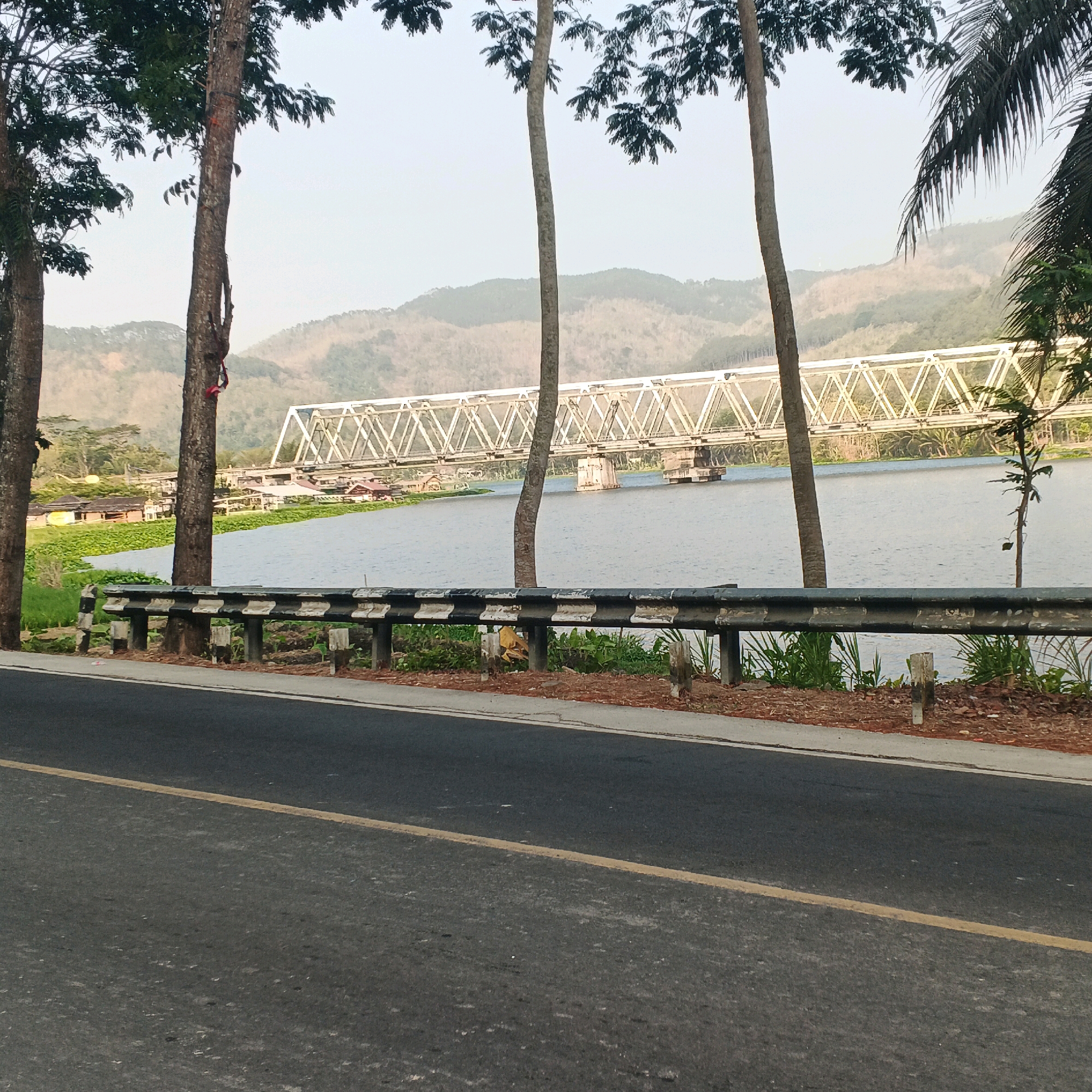 In Kebasen, Indonesia, a view captures a body of water with a railway bridge spanning across it. The bridge is constructed with a lattice-like metal structure supported by concrete piers. On the far bank of the water, a cluster of buildings with red roofs is visible, nestled amidst lush green vegetation. Beyond the buildings and water, rolling hills form the backdrop, their slopes covered in trees. In the foreground, a road runs parallel to the water, separated by a metal guardrail painted with alternating black and white stripes. Tall trees with dark green foliage stand between the road and the guardrail, their trunks partially obscuring the view of the bridge and the landscape beyond. The sky is overcast, casting a muted light over the entire scene.