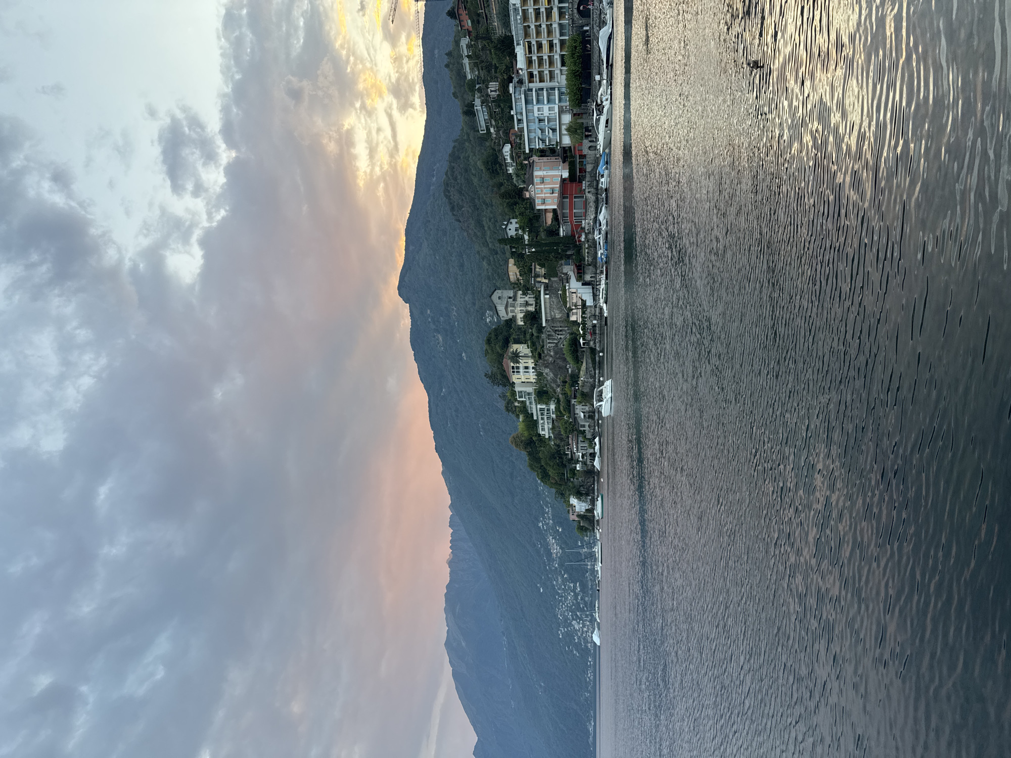 The image captures a serene waterfront scene in Ascona, Switzerland, during what appears to be late afternoon or early evening. The sky is filled with a dramatic display of clouds, with soft hues of pink and orange mingling with shades of grey and blue, suggesting either sunrise or sunset.
In the foreground, the calm surface of a body of water, likely Lake Maggiore, reflects the subdued light, with gentle ripples disturbing the otherwise smooth expanse. A few small boats are visible docked along the shore.
The midground features the picturesque town of Ascona, nestled along the hillside. Buildings of various architectural styles, from traditional to modern, are scattered across the verdant slopes. Some structures are painted in light colors, while others have more vibrant facades. The town is densely built, climbing up the mountain that forms the backdrop.
The background is dominated by a large, imposing mountain range, its dark green slopes partially obscured by the dusky light. The rugged peaks of the mountains rise majestically, framing the charming town. The overall atmosphere is one of tranquility and natural beauty, with a hint of urban charm. There are no people or animals clearly visible in the main scene, contributing to the peaceful ambiance.