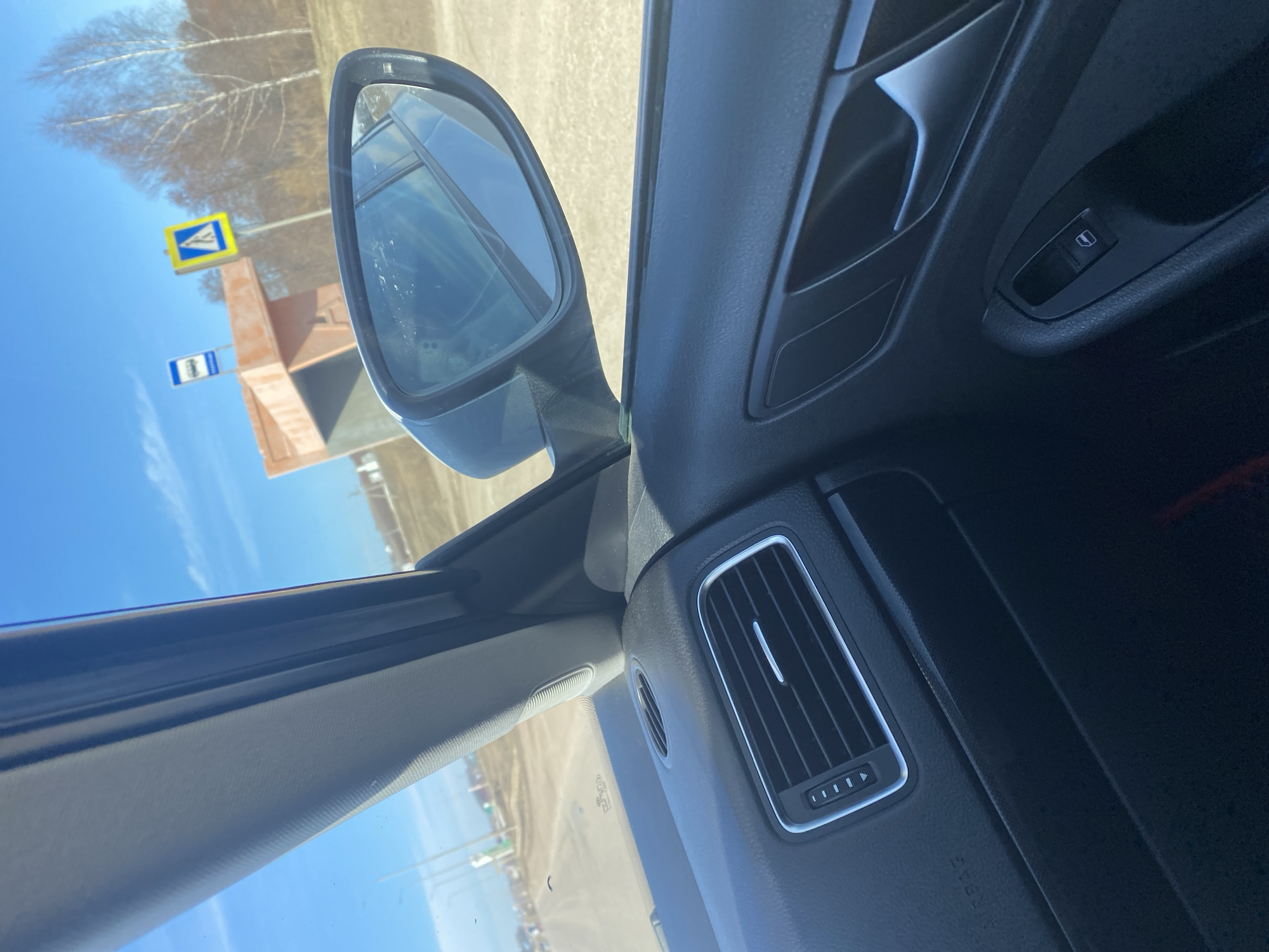 The image is taken from the interior of a vehicle, showing a side mirror reflecting a street scene. Visible in the mirror's reflection are road signs, buildings, and trees under a blue sky. The interior of the car features a window control, a vent, and the dashboard. The location is Verkhniye Tatyshly, Russia, as indicated by the bus stop sign written in Russian.