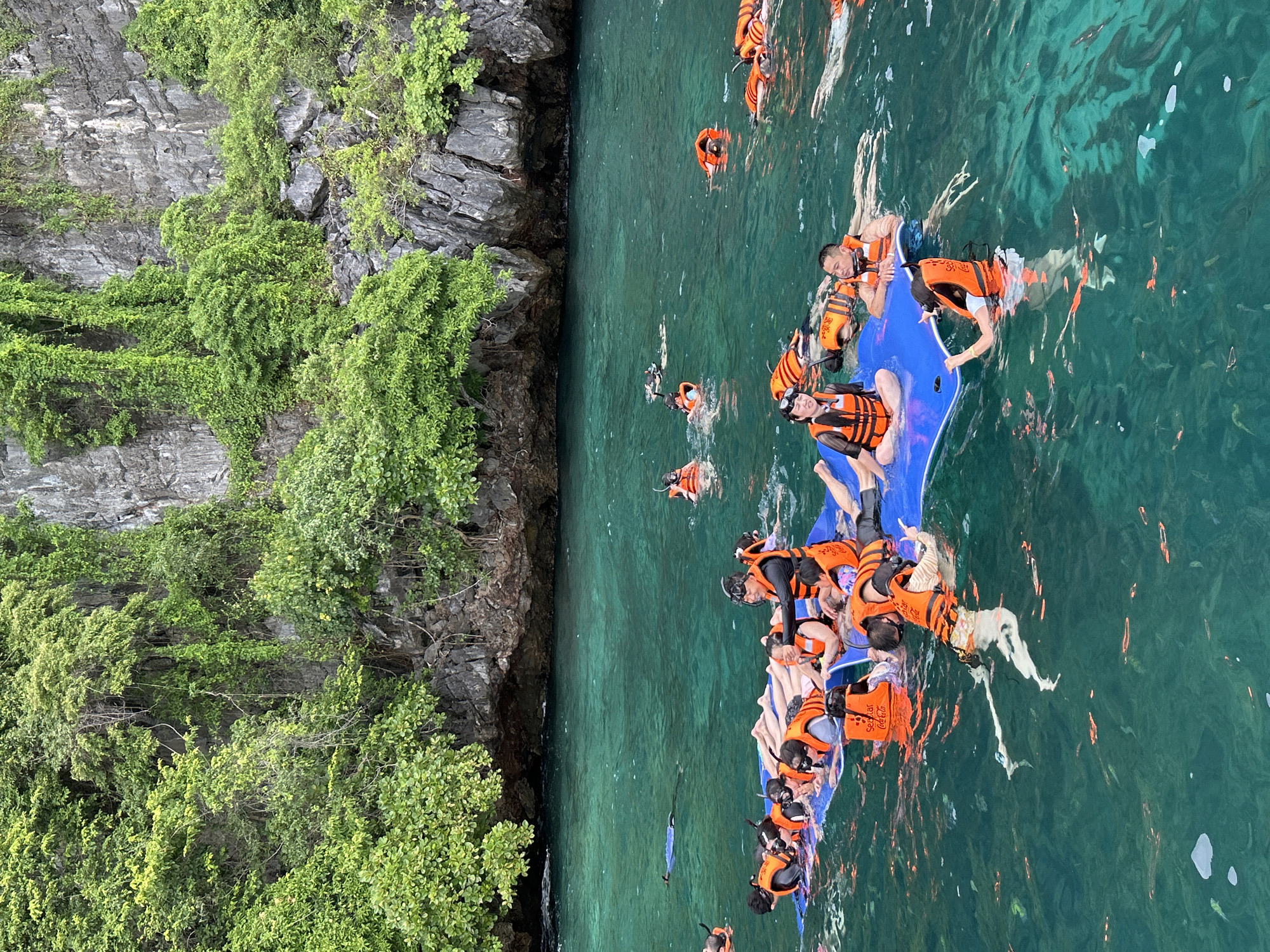 A group of tourists are floating on a blue raft in clear, turquoise water near a rocky cliff face covered in lush green foliage. Many of the individuals are wearing orange life vests and snorkeling gear, suggesting they are engaged in recreational water activities. Some are lying on the raft, while others are in the water, with several visible snorkeling. The scene appears to be a popular tourist spot in Ao Nang, Thailand, known for its stunning natural beauty and opportunities for island hopping and water sports. The water is clear enough to see some fish below the surface.