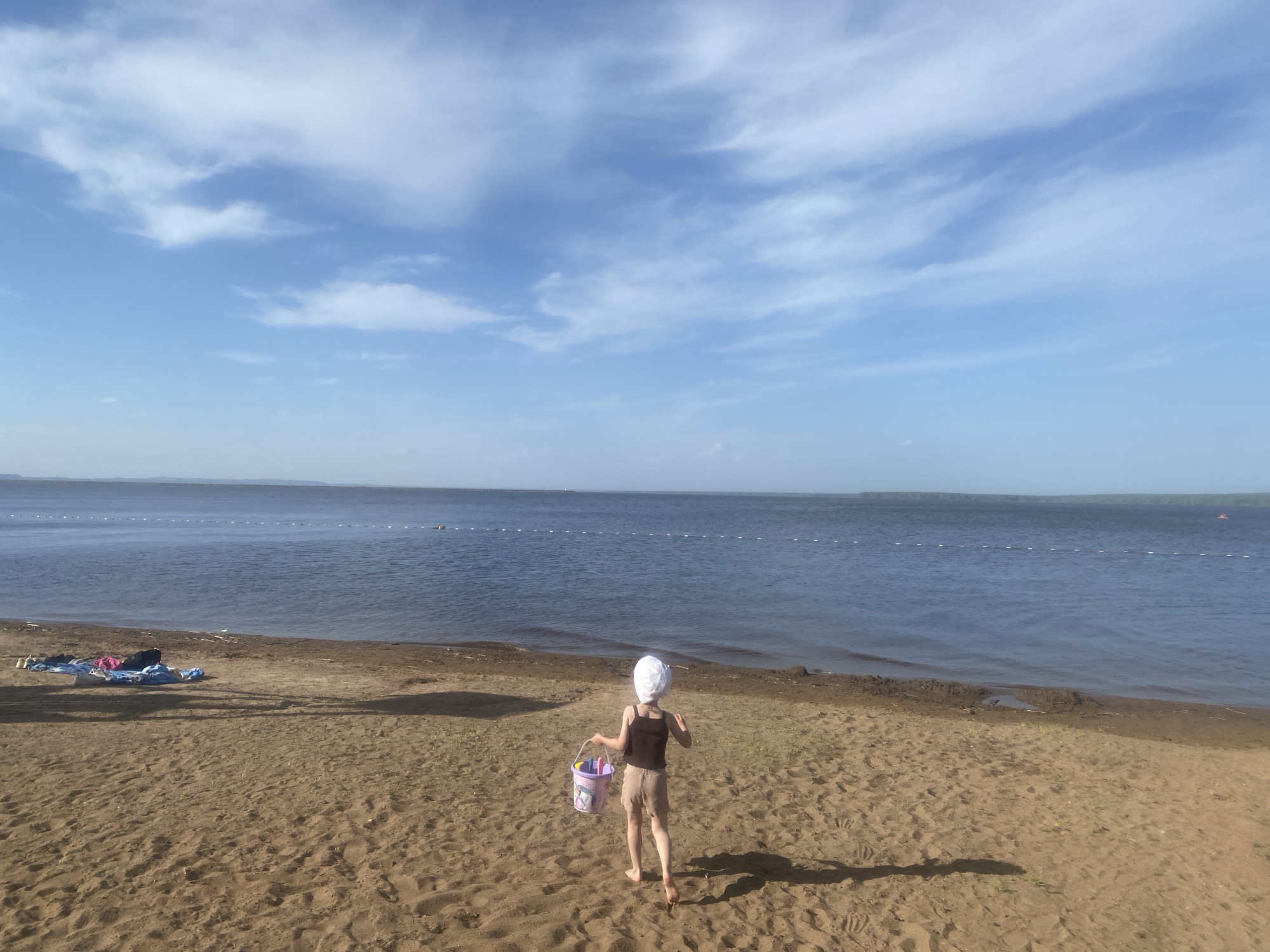 A child wearing a brown tank top, tan shorts, and a white hat walks away from the viewer, carrying a purple bucket. The child is barefoot on a sandy beach. In the background is a body of water, with a line of buoys. The sky is blue with scattered clouds. A pile of blankets and towels is visible to the left. The location appears to be a beach on the Kama River near Chaikovsky, Russia.