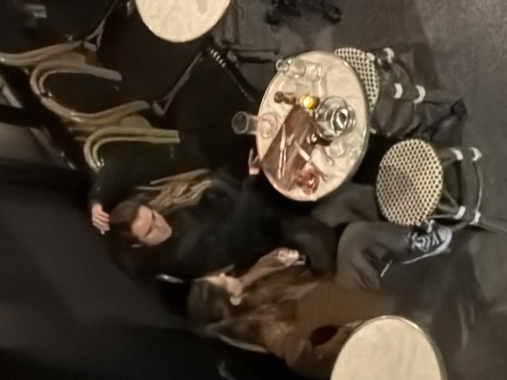This media file captures an overhead view of two individuals seated at a small round table in an outdoor café setting, likely at night.
**Subjects:** Two people are visible. The person on the left has long, light brown hair and is wearing a dark top, with their face partially visible. The person on the right is less distinct, dressed in dark clothing, with black and white sneakers visible.
**Objects:** A small, light-colored round table serves as the central focal point, displaying several clear glasses, a small bottle or carafe, and a small, glowing light source, possibly a candle or a decorative lamp. Surrounding the main table are other similar round tables and chairs with distinctive, possibly ornate, metal frames and patterned seat/backrests.
**Activity or event taking place:** The individuals appear to be relaxing and socializing, enjoying drinks at the café.
**Notable details:** The scene is set at night or in the evening, indicated by the dim lighting and the illuminated item on the table. The style of the tables and chairs suggests a classic European café ambiance, consistent with a Parisian street-side establishment. No specific weather conditions are discernible, and facial expressions are not clear enough to determine specific emotions, though the atmosphere appears relaxed.
**Visible text:** There is no visible text in the image.