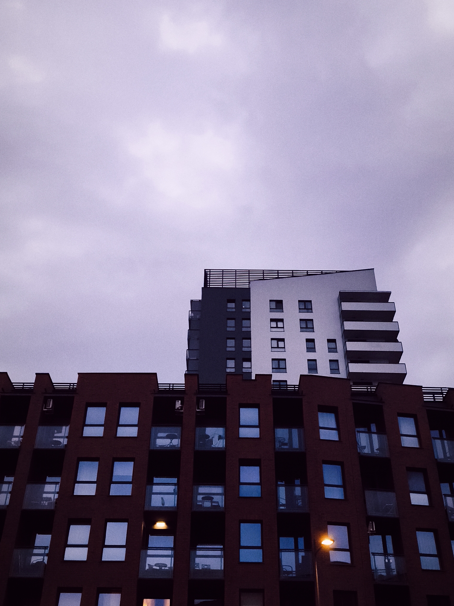 The image shows a contemporary apartment building with a distinctive red brick facade and numerous windows and balconies. Above this building, a modern white and gray building rises with a striking cantilevered section. The sky is overcast and has a purplish hue, suggesting it is either dawn or dusk. A single streetlamp is visible on the right side, emitting a warm glow, indicating it is likely evening. The architectural style suggests an urban setting in Gdańsk, Poland. The image captures a slice of urban life, focusing on the geometric forms of the buildings against the dramatic sky. No people are visible in the image.