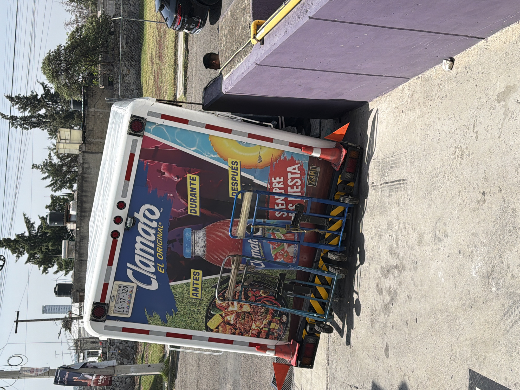 The scene captures a delivery truck parked near a purple-colored concrete structure. The truck is adorned with a prominent advertisement for Clamato, a tomato juice beverage, displaying images of various dishes, including seafood, alongside the phrases "ANTES," "DURANTE," and "DESPUÉS." A blue hand truck is positioned behind the vehicle. The license plate on the truck reads "LG-07-329." Adjacent to the truck, a person is observed in the background. The surrounding environment is partially urban, with trees, power lines, and buildings visible in the distance. The presence of Mexican license plates and Spanish text on the truck's advertisement suggests the location is in Mexico. The image's metadata confirms it was taken in San Bernardino Tlaxcalancingo, Mexico.