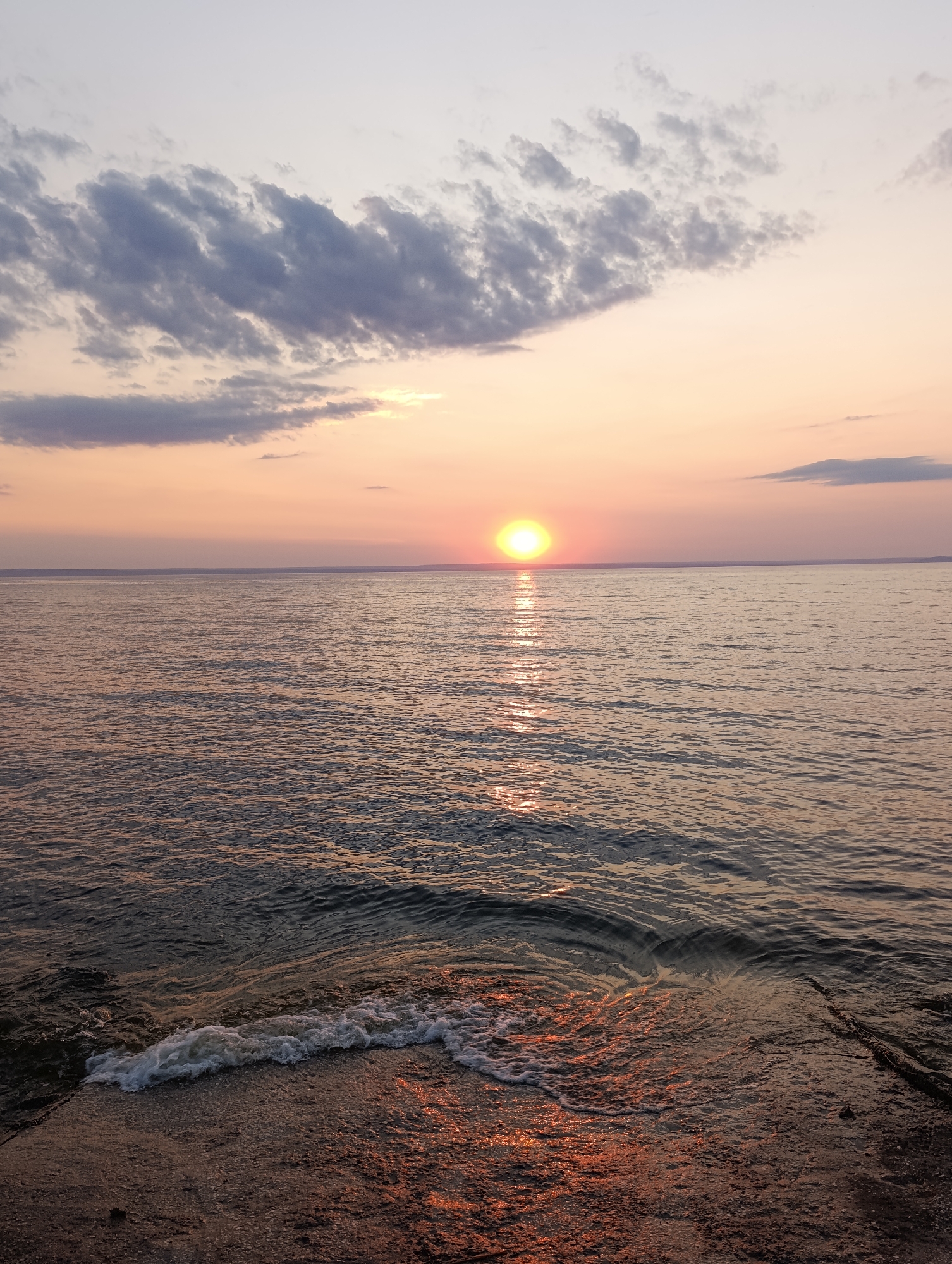 This is a vertical, eye-level, wide shot of a sunset over a body of water. The sun is a bright, yellow-orange orb situated in the center of the frame, low in the sky. The sky transitions from a pale lavender at the top to a soft peach and pink hue near the horizon. Wispy, dark gray clouds are scattered across the upper portion of the sky. The water is a deep, dark blue with subtle ripples reflecting the colors of the sunset. In the foreground, a concrete structure juts into the water, with a small wave breaking against it, creating white foam. The concrete appears rough and textured, with hints of reddish-brown from the sunset's reflection. The scene is tranquil and serene, with no people or animals visible. The location is unknown.