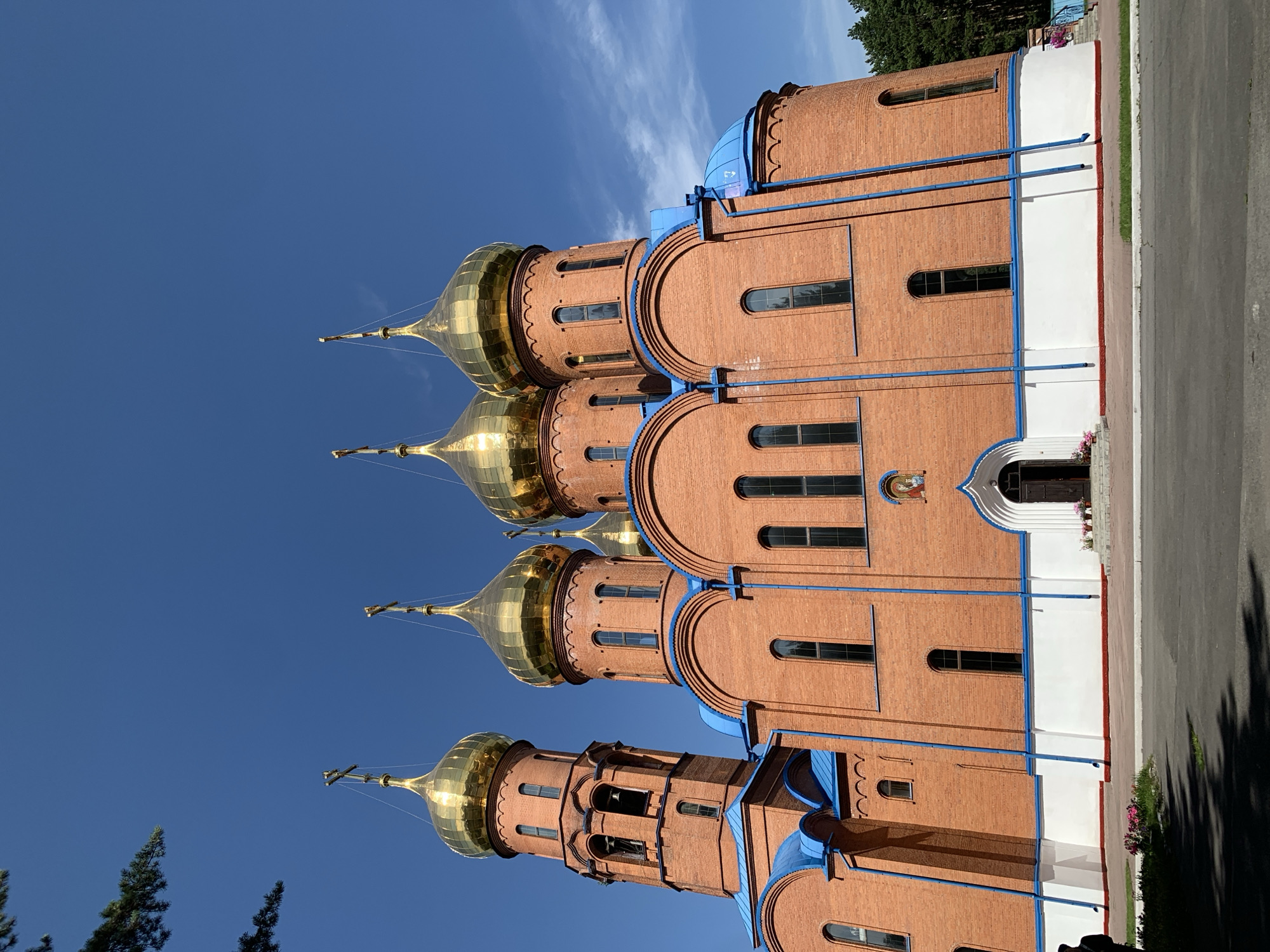 The image shows the exterior of an Orthodox church with multiple golden domes and a red brick facade. The church has a tall bell tower to the left, also with a golden dome topped by a cross. The domes and rooflines are adorned with blue accents. The church is set against a clear blue sky with some wispy clouds. In the foreground, there is a paved area with some greenery and flowers. To the right, a line of pine trees is visible. The church is the Holy Trinity Cathedral in Prokopyevsk, Russia.
