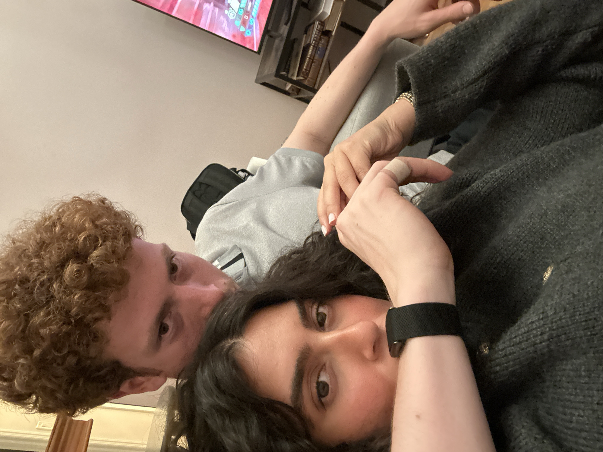 A couple is pictured indoors, likely in a residential setting. A male with curly red hair is lying on his back, with a female, with dark hair, lying on top of him. The female is looking directly into the camera. Both appear to be relaxing or resting. There is a television visible in the background. The location is likely somewhere in the United States of America and possibly New York, based on the prompt.