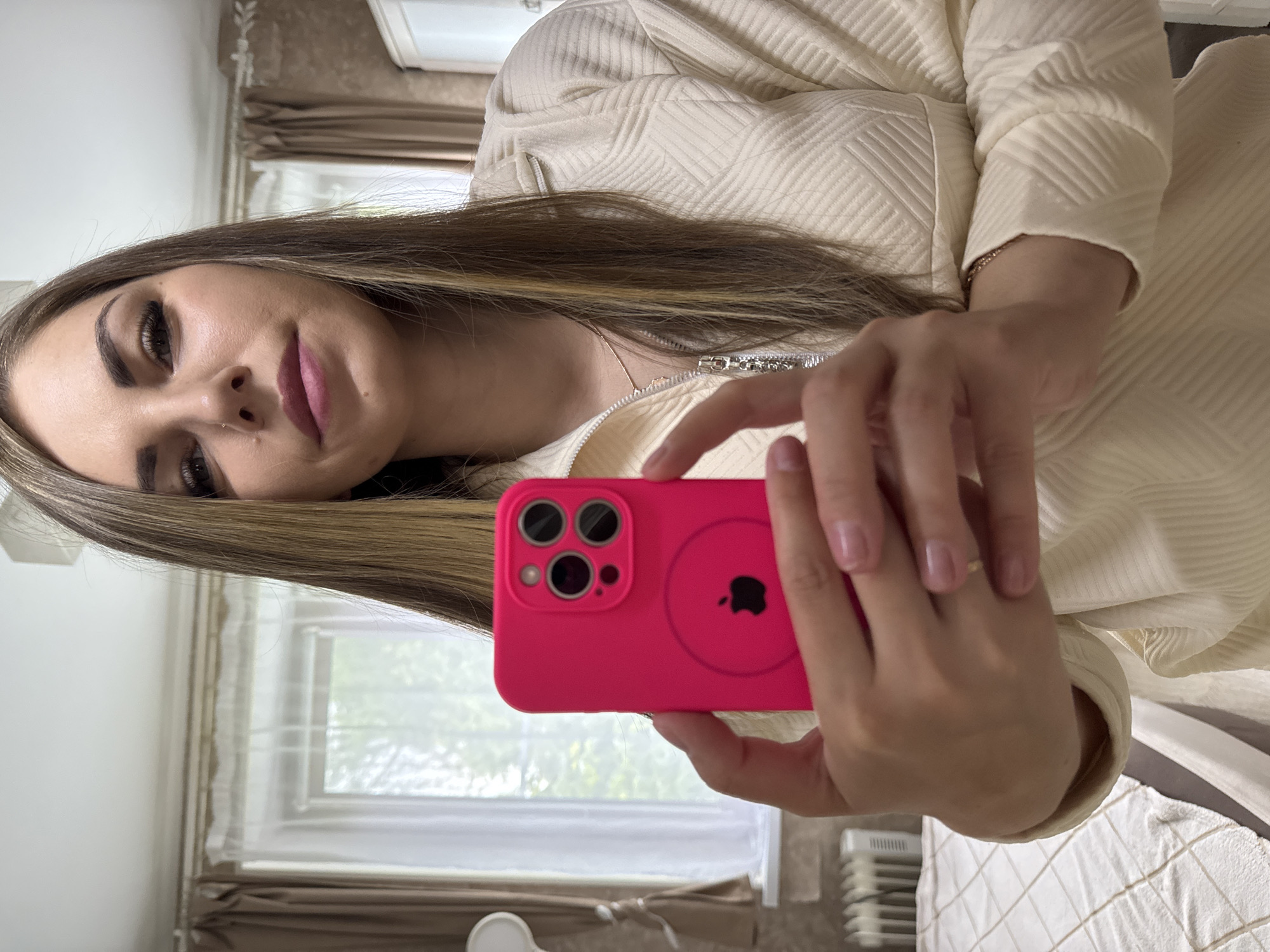 A woman holds a pink iPhone with a MagSafe attachment in front of her, possibly taking a selfie. She has long brown hair with blonde highlights and is wearing a light-colored jacket. The background includes a window with sheer curtains, a white wall, and what appears to be a radiator. It is not possible to determine if this image was taken in Раздолье, Russia.