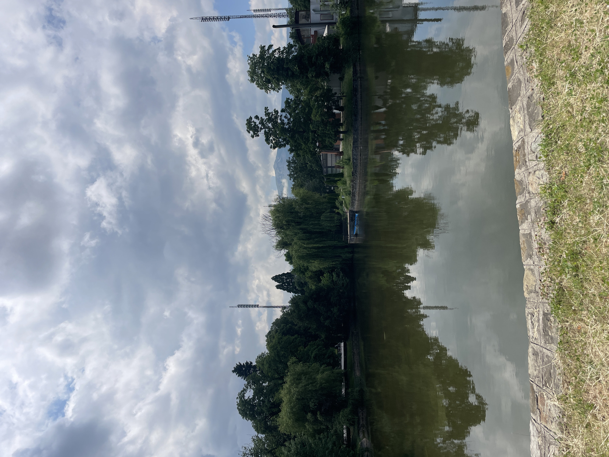This landscape photograph captures a tranquil waterside scene under a partly cloudy sky, likely in Sofia, Bulgaria. The image is dominated by a calm body of water, possibly a lake or canal, which acts as a mirror, reflecting the sky and the opposing bank with remarkable clarity.
On the immediate right side of the frame, a strip of dry, light green grass transitions into a stone or concrete retaining wall that runs along the water's edge. The texture of the wall is visible, made of irregular stone blocks.
The opposite bank is lush with a variety of green trees, including a prominent weeping willow with branches gracefully extending towards the water. Behind the canopy of trees, several buildings are partially visible, hinting at an urban or suburban setting adjacent to the natural area. In the background, a large mountain range, likely Vitosha mountain, rises under the clouds, partially obscured by atmospheric haze.
Above the tree line on the far bank, two tall, slender metal structures are visible. These appear to be lighting towers, possibly for a sports complex or stadium, with one more centrally located and the other further to the right. Near the weeping willow, a small blue object, possibly a boat or a section of a dock, can be discerned on the far bank.
The sky is filled with expansive, soft white and grey clouds, interspersed with patches of clear blue sky, suggesting a bright, overcast day. The reflections in the water are vivid, duplicating the cloudy sky, the silhouettes of trees, and the tall structures.
The photograph depicts a static, peaceful daytime scene. There are no people visible, and no specific activities are taking place, contributing to the quiet ambiance of the natural and semi-urban environment. No visible text is present in the image.