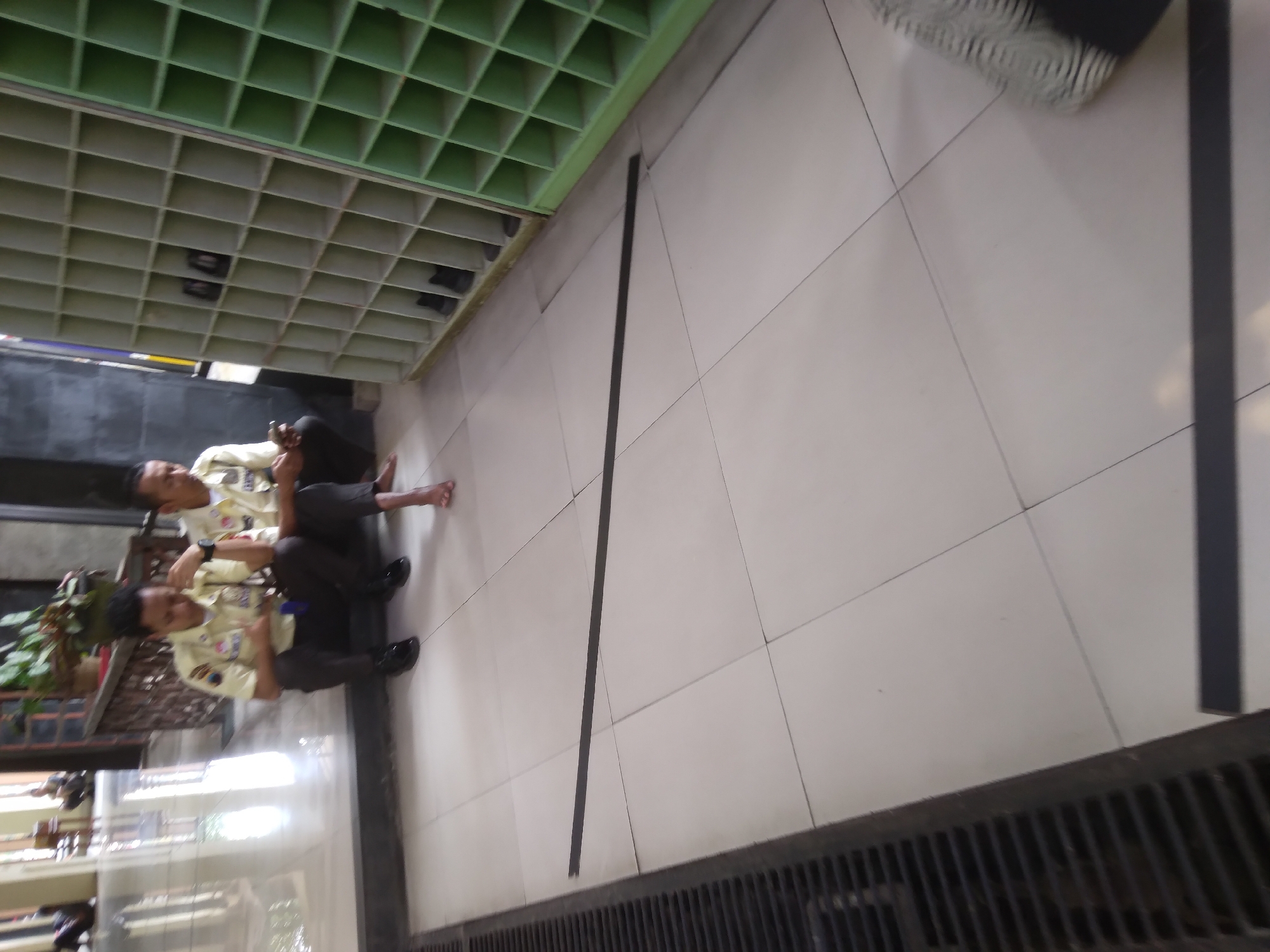 Two individuals, appearing to be male, are seated on the floor in front of a shoe rack. They are wearing matching light-colored uniforms with dark pants. One individual is holding a mobile phone and appears to be using it. The other individual is looking in the same direction. The shoe rack is large and has many compartments, some of which contain shoes. The floor is tiled with a light-colored material. There is a black line on the floor in front of the individuals. To the left, there is a drainage grate. The scene appears to be indoors, possibly a public building or facility. The overall lighting suggests it is daytime. This scene is located in Desa Pamijen, Kecamatan Sokaraja, Indonesia.