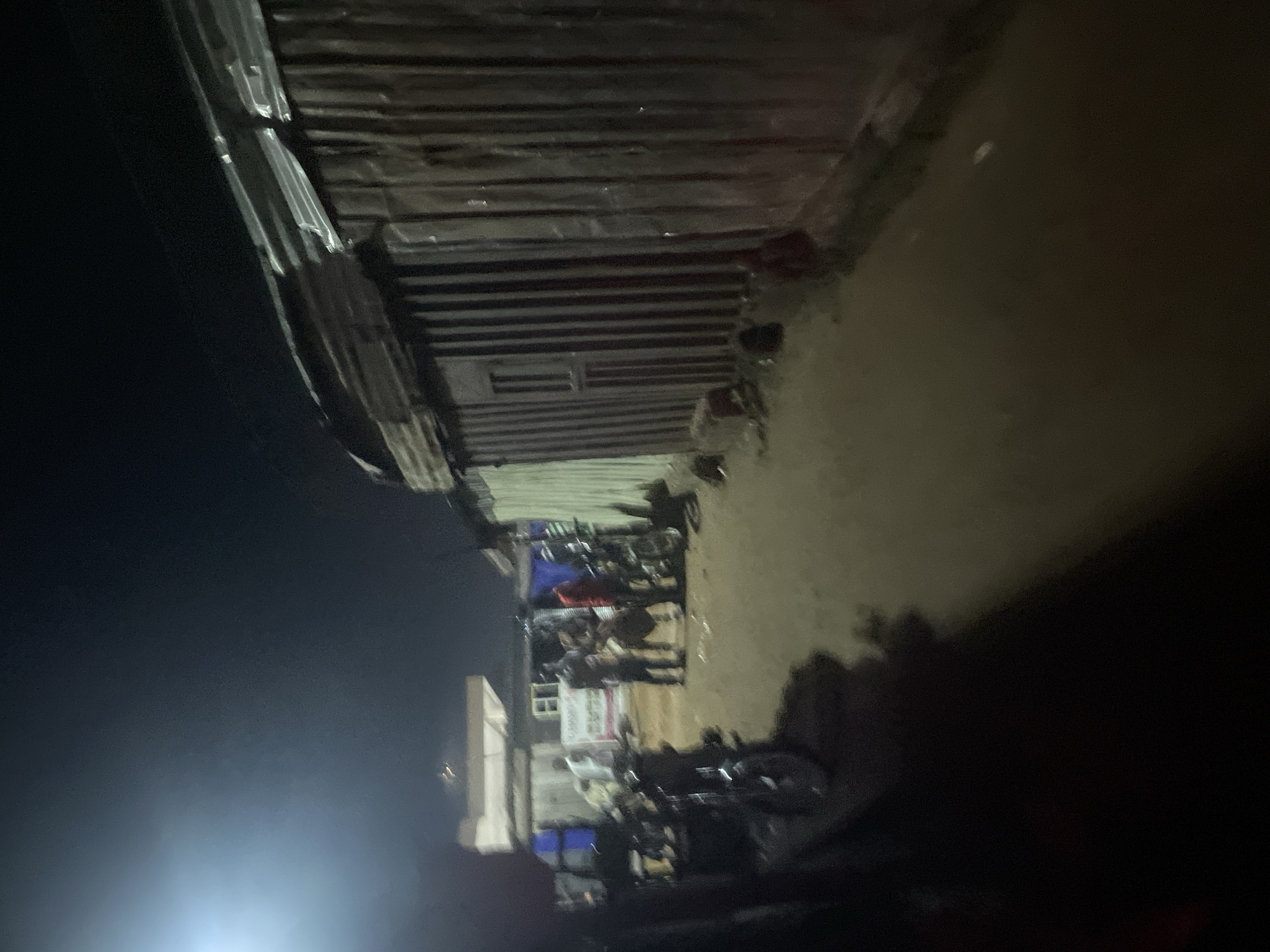 The image depicts a dimly lit outdoor scene, likely in a residential or commercial area of Lokoja, Nigeria, at night. The foreground is dominated by dark shadows, obscuring details. In the midground, several individuals are gathered. One person, wearing a red shirt, stands centrally. To their left, another person in a dark shirt appears to be speaking or interacting with a group. Further left, a person in a white garment is visible near a building. There are several motorcycles parked in the scene, indicating a mode of transportation. The background features buildings constructed from corrugated metal, characteristic of informal settlements or makeshift structures. One building has a visible window and a corrugated metal roof. The ground appears to be unpaved, with some scattered debris. The overall atmosphere is somewhat chaotic and appears to be a community gathering or street scene after dark.