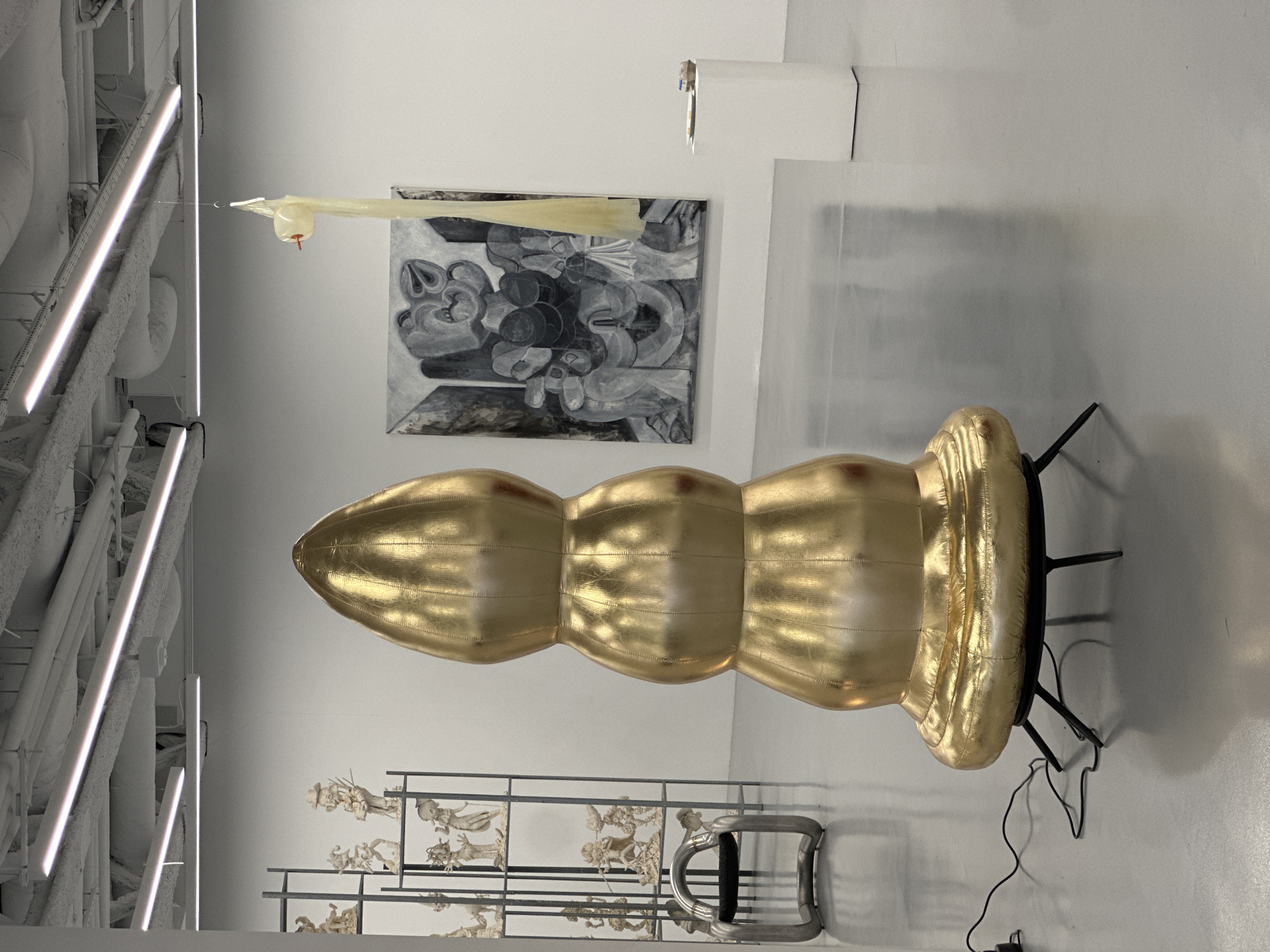 The image shows an art installation in a gallery, possibly located in London, United Kingdom, given the prevalence of such spaces in the city. The environment is a well-lit, white-walled gallery space. Dominating the scene is a large, gold-colored, inflatable sculpture resembling a stylized, segmented form. This object rests on a black metal stand.
Behind the sculpture, mounted on the wall, is a black and white abstract painting. Suspended from the ceiling is a clear plastic tube with a small, possibly deflated, colored item attached. Below the sculpture is a metal shelving unit displaying various small, white figures.
An array of white fluorescent lights is visible on the left side of the frame, mounted on the ceiling. A white box is visible on the wall, possibly an electrical box. The floor is a light gray color. There are electrical wires snaking from the base of the gold sculpture across the floor. No people are visible in the image.
