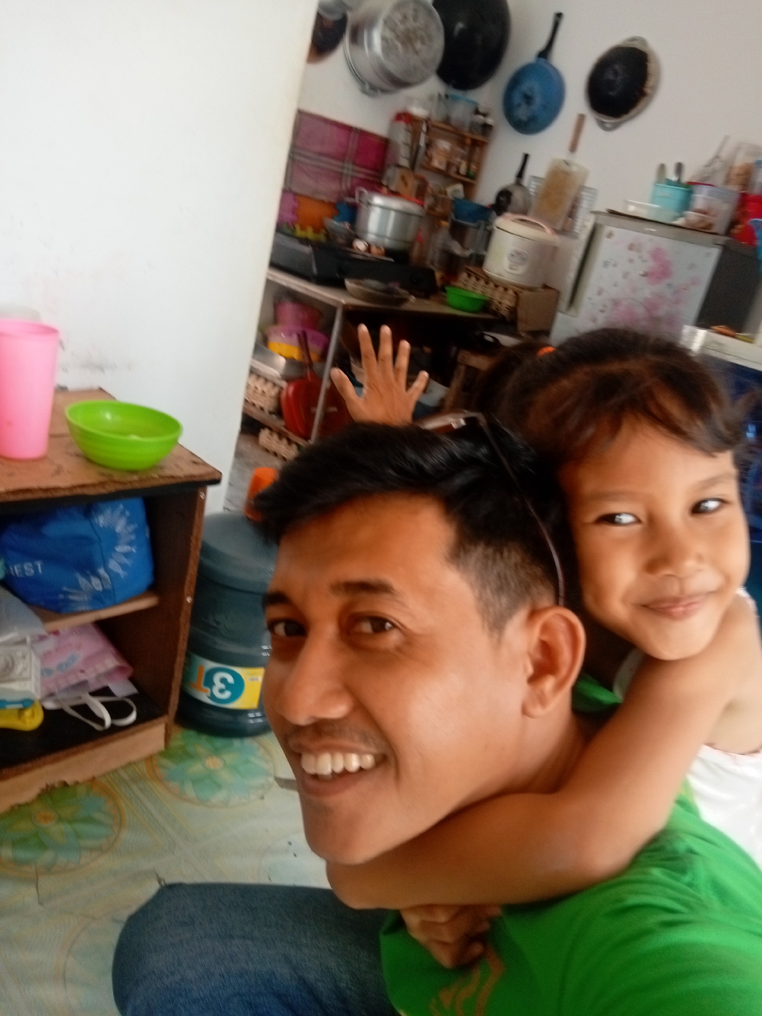 A young girl with dark hair, her arms around a man's neck, smiles at the camera. The man, who appears to be her father, has dark hair and a mustache and is also looking at the camera. They are likely in their home in Batam, Indonesia, as evidenced by the presence of a water dispenser with a "3" logo, a common sight in Indonesian households. The background shows kitchen items like pots and pans hanging on the wall, shelves with various containers and appliances, and a pink cup and green bowl on a wooden shelf. The floor has a patterned tile design.