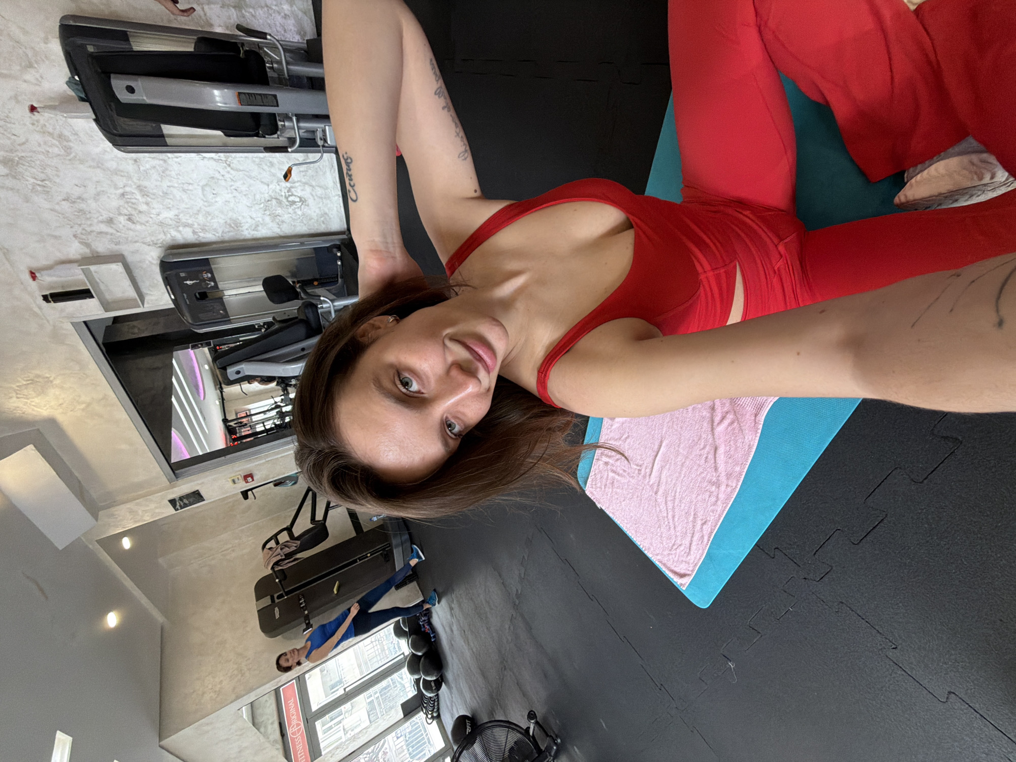 The image displays the interior of a gym, identified as Fitness Original, located in Budva, Montenegro. The central figure is a woman with brown hair and green eyes, wearing a red athletic top and matching red pants with a hip cutout, seated on a turquoise yoga mat with a pink towel. She is taking a selfie, holding the camera with her left hand, and her right hand is placed behind her head. Tattoos are visible on her right forearm, reading "Семья" and "this too shall pass." The gym floor consists of interlocking black rubber mats. In the background, an air bike is partially visible on the left, and a stack of black kettlebells is positioned near a large window. Another woman, dressed in a blue top and dark leggings, stands farther back next to a grey and black selectorized weight machine. Additional gym equipment and a mirrored wall are present. Light-colored, textured walls are visible, and white shelves holding cleaning bottles are mounted on the wall. A glass door reflecting "FITNESS ORIGINAL" provides a view of an urban exterior.
