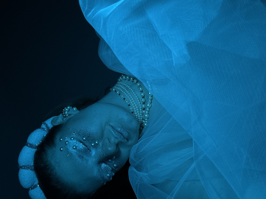 A close-up, forensically lit portrait captures a female individual under dominant cool blue lighting. Her head is tilted, with her eyes appearing closed or heavily shadowed. The subject wears a white, ribbed headband with shimmering accents. Her face is adorned with numerous small, spherical white beads, concentrated around her eyes, nose, and cheeks, alongside white glitter or eyeshadow on her eyelids. A multi-strand pearl necklace, consisting of approximately six layers, encircles her neck. A cluster of small white beads is visible on her right earlobe, serving as an earring. Her body, partially visible, is draped in a voluminous amount of sheer, light blue fabric, resembling tulle or chiffon, which fills the lower and right portions of the frame. The background is uniformly dark, offering no discernible features. The subject is posed in a serene or contemplative manner. No specific location within Kazan, Russia, can be identified from the visual evidence in this image.