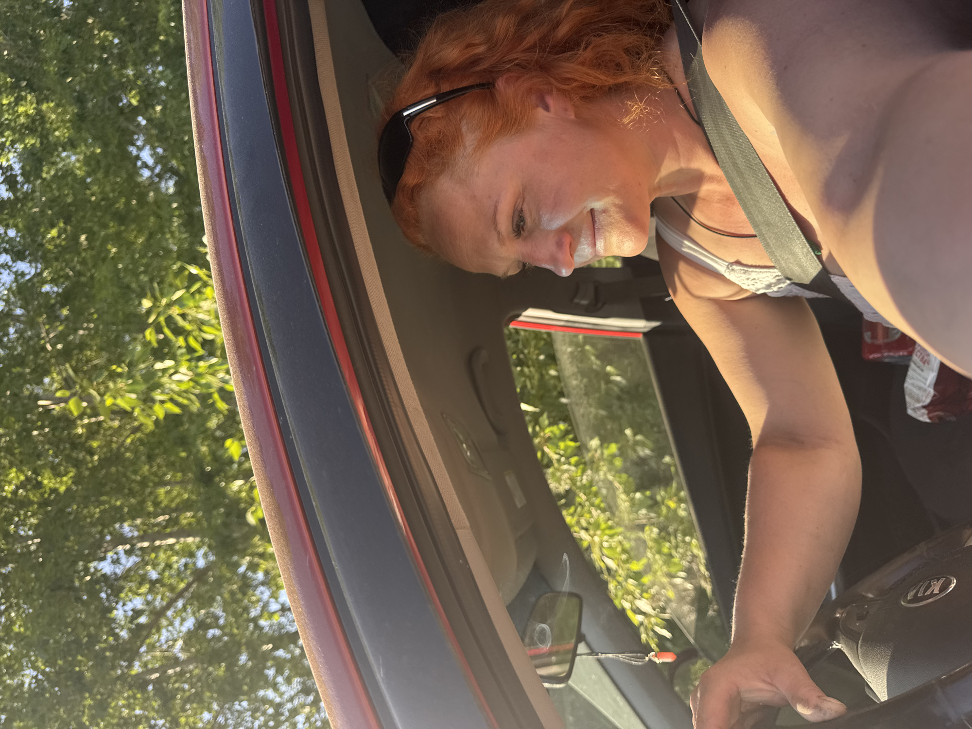 A red-haired woman is leaning into a red Kia car. She is wearing a seatbelt, a black headband, and a white top with lace. Her left hand is on the steering wheel. Part of her face is visible, with a slight smile. Outside the car, there is a view of green trees against a partly sunny sky. The location is the city of Chaikovsky, Russia. The car is parked. There is a rearview mirror hanging by the windshield.
