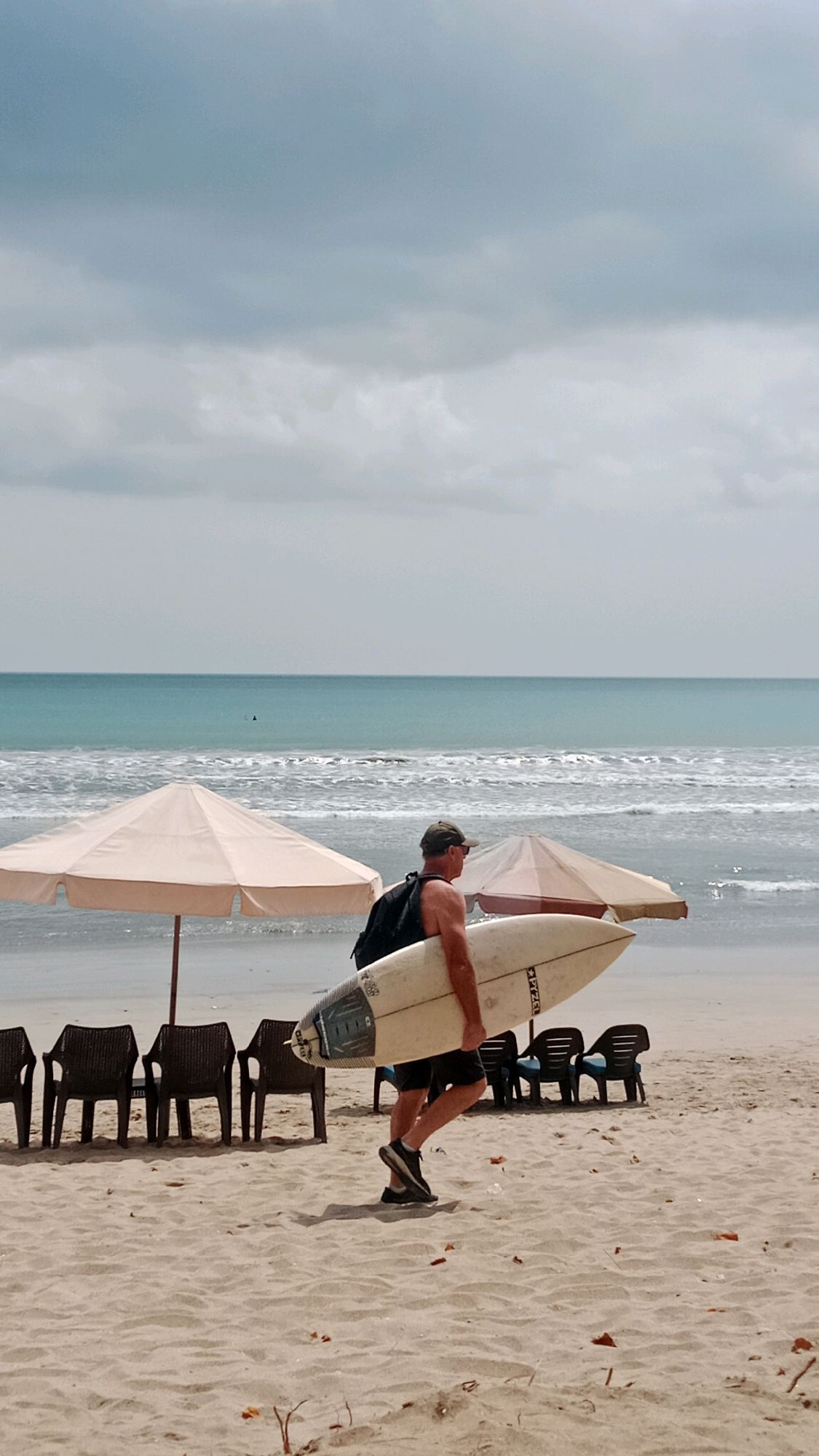 This image captures a man walking along the sandy beach of Kuta, Indonesia, carrying a white surfboard under his arm. He is wearing a dark backpack, black shorts, and black sneakers. The man is walking from left to right, with his gaze directed forward. In the background, the turquoise ocean meets the horizon under a cloudy sky. Several beach chairs are visible in the middle ground, along with two large beach umbrellas. The foreground is dominated by the sand, with scattered dry leaves and debris. The scene depicts a typical beach day with a surfer heading towards or away from the water.