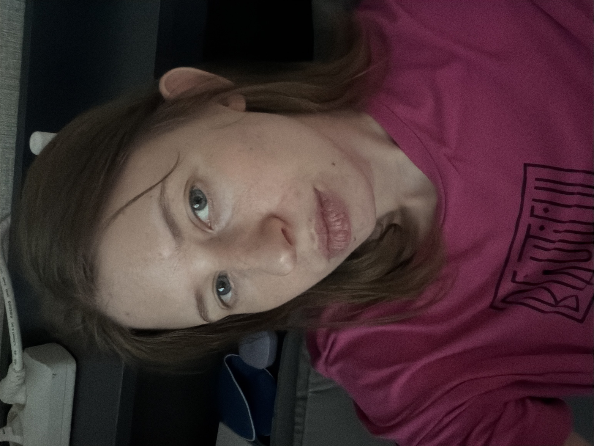 A young woman with light brown hair and blue-green eyes is looking directly at the camera. She is wearing a pink t-shirt with a black graphic design on the front. Her head is tilted slightly to her right, and her mouth is slightly pursed. The background is dark and out of focus, but appears to be indoors, possibly a bedroom. There is a power strip and some pillows visible in the upper left and mid-left of the image, respectively. The overall lighting is dim.