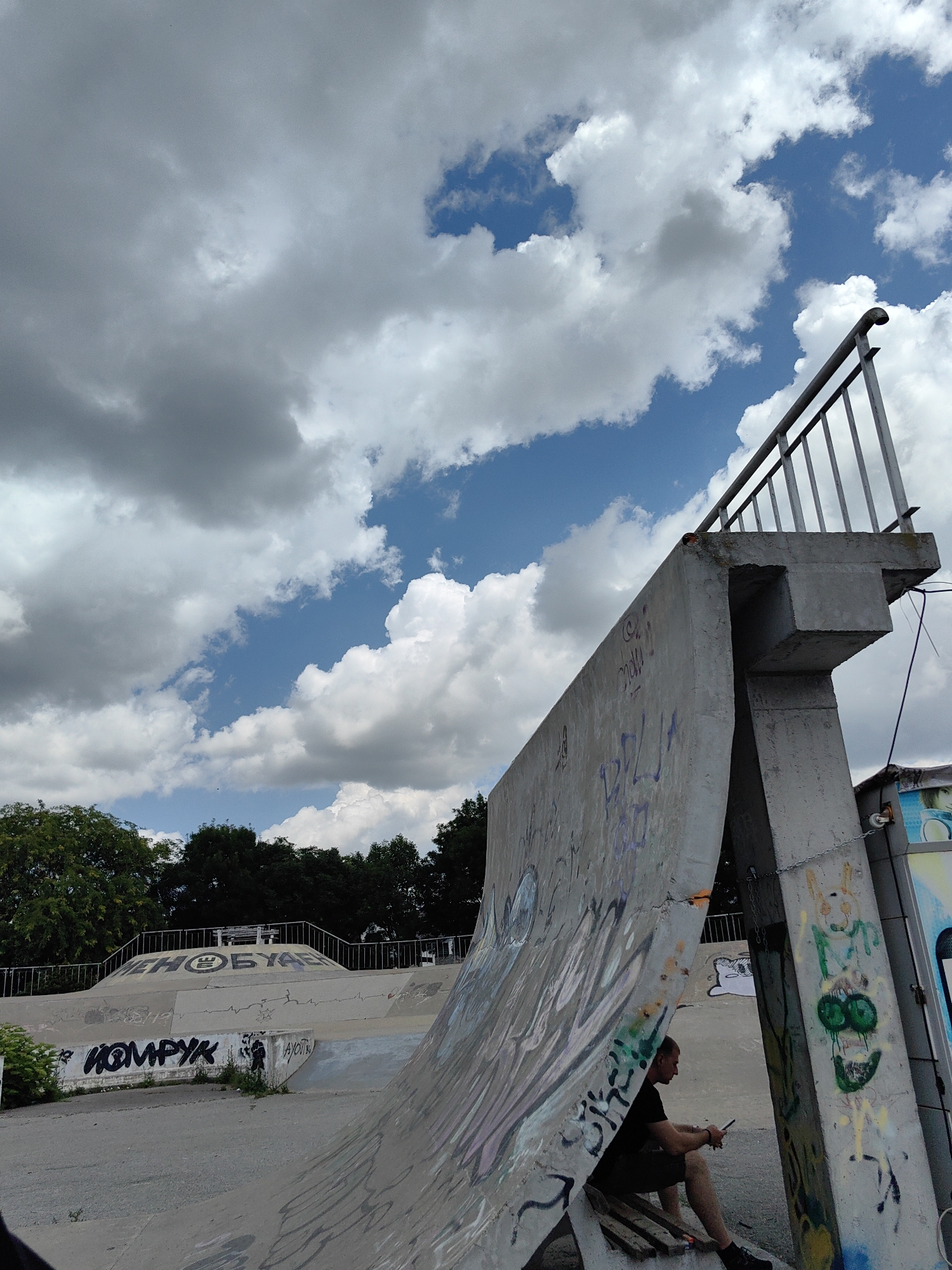 The image depicts a concrete skate park under a cloudy sky. The foreground features a large, curved concrete ramp covered in graffiti, with a person sitting on a wooden bench underneath an overhang, looking at their phone. In the background, more skate park structures are visible, along with trees and a railing. The sky is filled with a mix of dark and light grey clouds, with patches of blue sky peeking through. The overall impression is of a casual, possibly late afternoon scene in an urban or semi-urban setting.
Visible text on the skate park structures includes: "КОМРУК" and some unreadable Cyrillic lettering, as well as graffiti tags and drawings. The setting appears to be a public park or recreational area. The person's posture suggests they are relaxing or waiting. The weather appears to be partly cloudy, possibly a bit overcast. No specific time of day is clearly indicated, but the lighting suggests it's daytime.