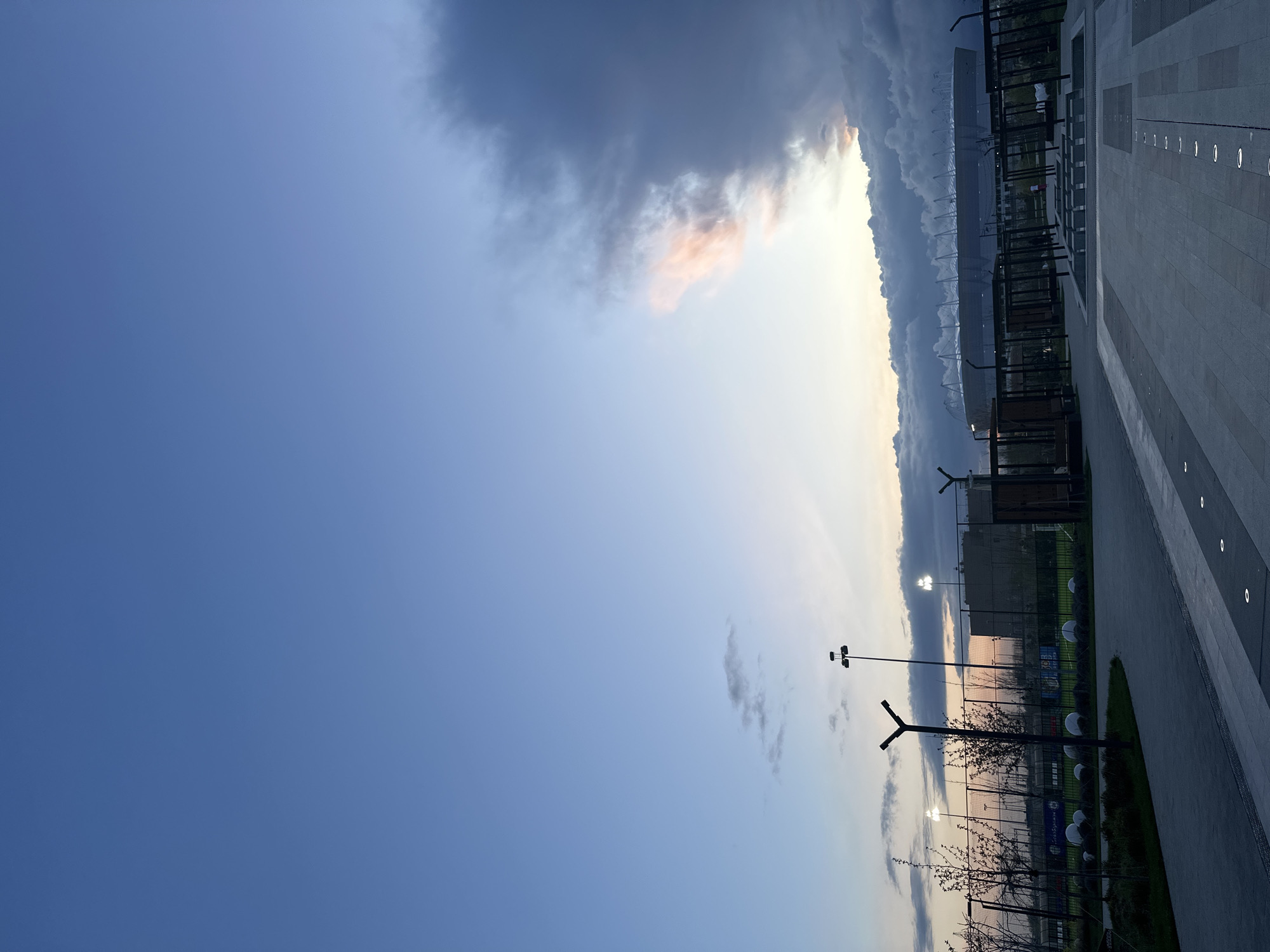 The image depicts a view in Rostov-on-Don, Russia, showcasing a modern paved area with integrated lighting. Visible on the right side is a portion of a building, possibly a stadium or arena, characterized by a metallic framework. A row of stylized lampposts is aligned along what appears to be a sports field or recreational area fenced off with netting. The sky transitions from a clear blue to a cloudy twilight or dawn with hues of pink and dark gray, indicating either sunrise or sunset. The overall scene suggests a contemporary urban landscape, possibly near a major sporting venue.