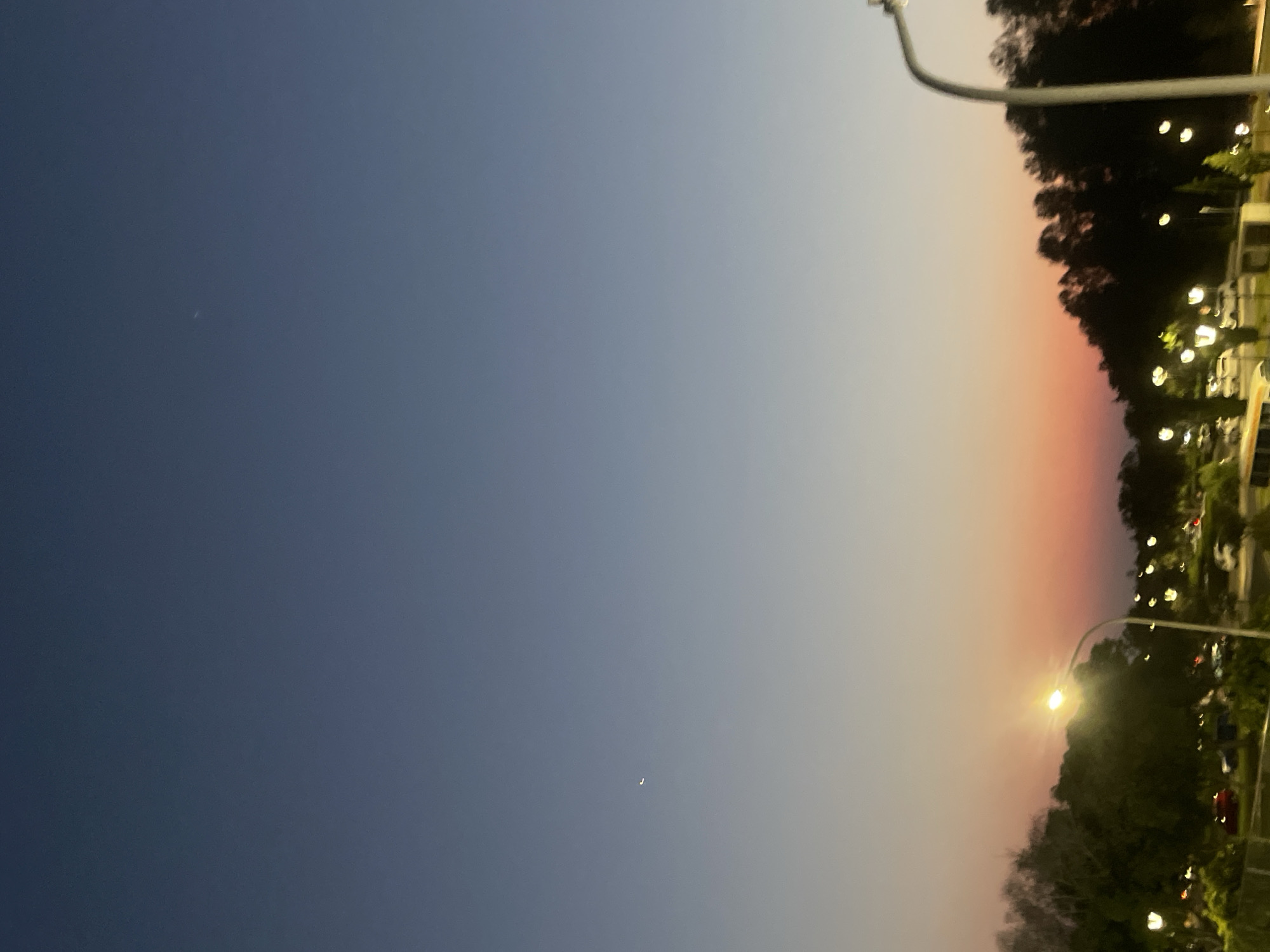 The image captures a twilight scene over a landscape that appears to be a public space or park area within Paphos Municipality, Cyprus. The sky transitions from a deep blue at the top to a pale gradient near the horizon, with hues of pink and orange indicating the setting or rising sun. A small, crescent moon is visible in the upper left quadrant of the sky.
In the foreground and midground, a line of trees forms a silhouetted backdrop against the colorful sky. Various lampposts with illuminated bulbs dot the landscape, suggesting an urban or semi-urban environment. Some of these lights appear to be streetlights, while others might be park or pathway lighting. There are also glimpses of parked vehicles, including what appears to be a white van or bus on the left, and several other cars further back, partially obscured by the foliage and lighting. The overall impression is of a peaceful evening setting, likely in a developed area of Paphos. No people are clearly visible in the image.