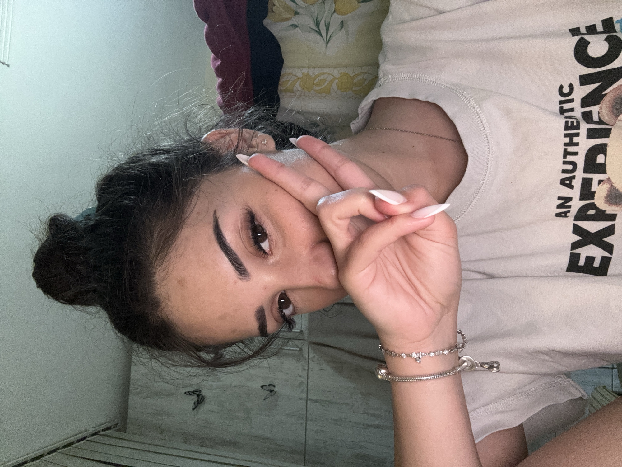 The image shows a close-up, selfie-style photograph of a young woman with her hair tied up in a messy bun. She is wearing a light-colored t-shirt with text and a graphic that is partially obscured. Her right hand is raised to her face, with two fingers extended in a peace sign. She is wearing a silver bracelet with charms on her left wrist and a delicate silver necklace. The background is indistinct but appears to be an indoor setting with a wall adorned with butterfly decals and some bedding or clothing in the upper right corner. The lighting suggests an indoor environment, possibly a bedroom. The image does not contain any elements that would allow for identification of its location within Plovdiv, Bulgaria.