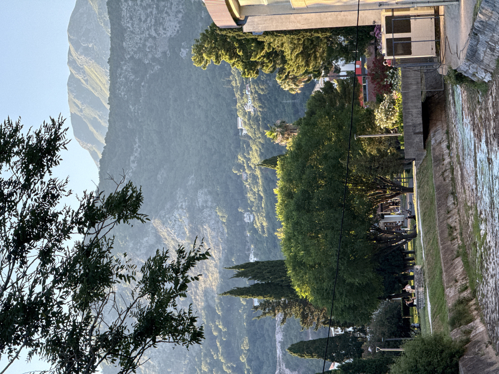 This image captures a view of the Budva coast in Montenegro. In the foreground, a dried-up riverbed with stone lining runs diagonally across the frame. On the right side of the riverbed, a modern building with a curved section is visible, adjacent to a white utility box enclosed by a metal fence. Across the riverbed, a park area is situated with several large trees, including a prominent, lush green tree in the center, and two tall, slender cypress trees. People are seen relaxing on benches in the park. In the background, the lush green slopes of mountains rise, dotted with white buildings. The sky is clear and blue.