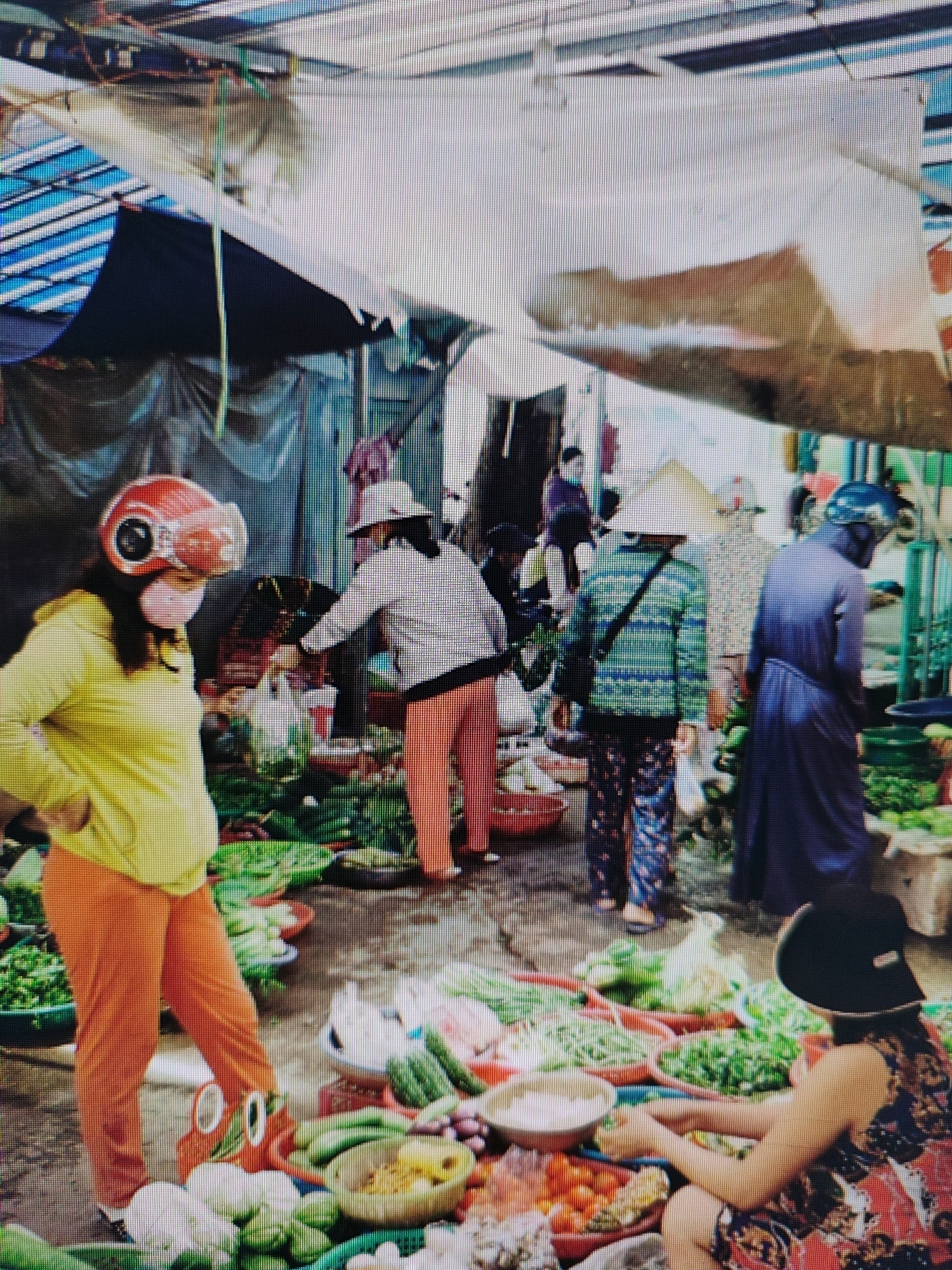 This image captures a bustling market scene. In the foreground, a person wearing a yellow long-sleeved shirt and bright orange pants stands with their hands on their hips. They are wearing a red helmet with a visor and a pink mask. To their right, a person in a conical hat and a blue and green striped shirt is visible, facing away from the camera. Further into the market, another person in a purple robe is seen walking away. A woman in a patterned dress and a black hat is seated in the lower right corner, holding a bowl. The ground is covered with various fresh produce, including leafy greens, cucumbers, and what appear to be tomatoes and onions, arranged in baskets and piles. The market is covered by a corrugated metal roof with some tarpaulin sections, creating a shaded environment. The overall impression is of a vibrant and active marketplace, potentially in Garut, Indonesia, given the style of hats and attire.