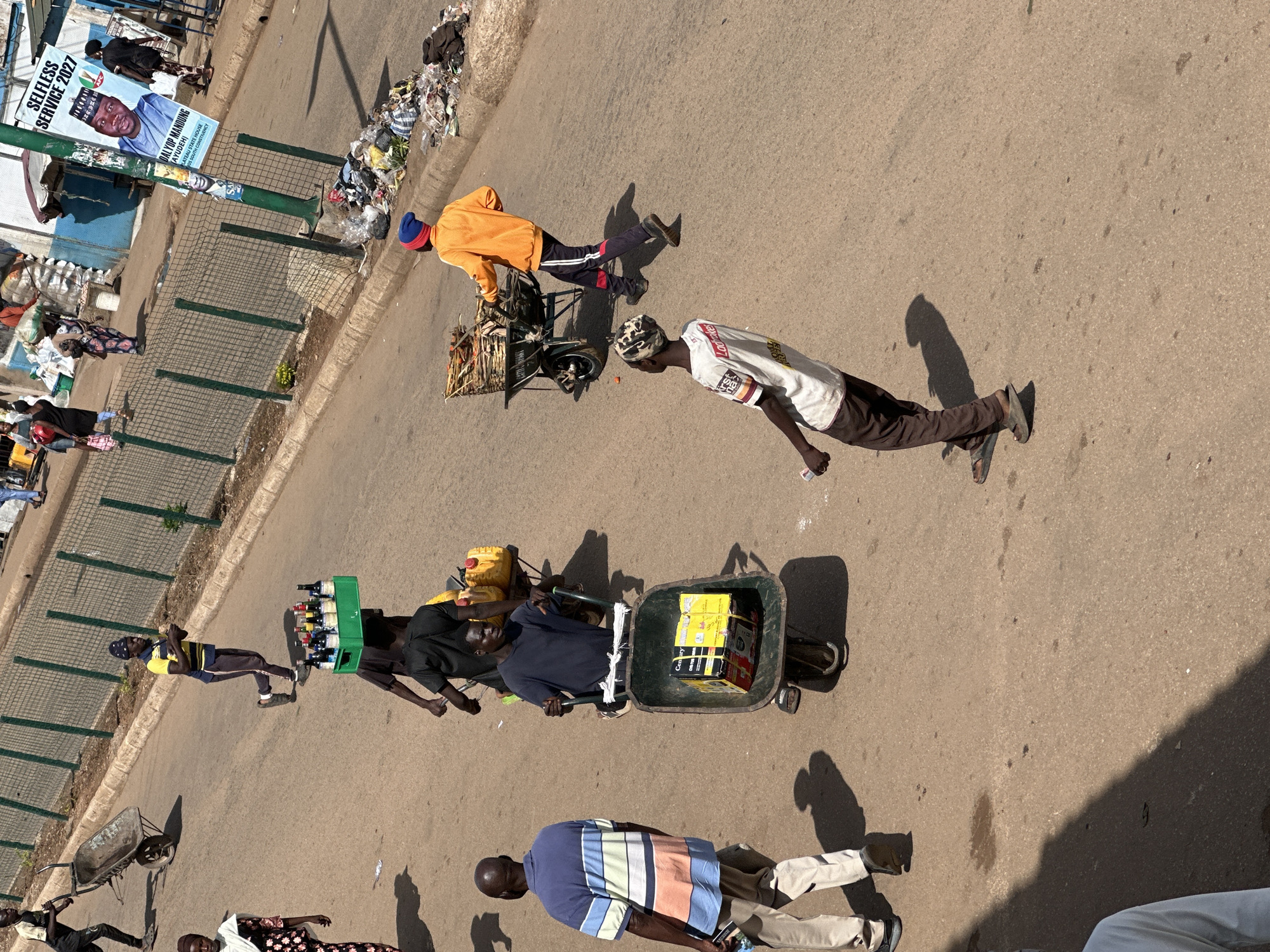 The image depicts a street scene in Bukuru, Nigeria, under bright daylight with distinct shadows. Several individuals are engaged in various activities. In the foreground, a man wearing a blue and white striped polo shirt and beige pants walks away from the camera. To his right, another man is pushing a wheelbarrow containing two yellow jerry cans and a black trash bag. Further back, a man in a dark t-shirt pushes a wheelbarrow with a large box inside, labeled "CENTURY PLUS". To his left, another man is carrying a green crate full of bottles on his head, walking alongside a person in a black shirt. On the left side of the image, a woman in a patterned dress is walking away. In the background, there are more people, some carrying loads, and a street lined with a green metal fence. A prominent poster on a lamppost displays an image of a man with the text "SELFLESS SERVICE 2027" and political party information. The ground is paved asphalt, with some scattered debris and small puddles. The overall environment suggests a busy market or thoroughfare with a mix of commerce and daily life.