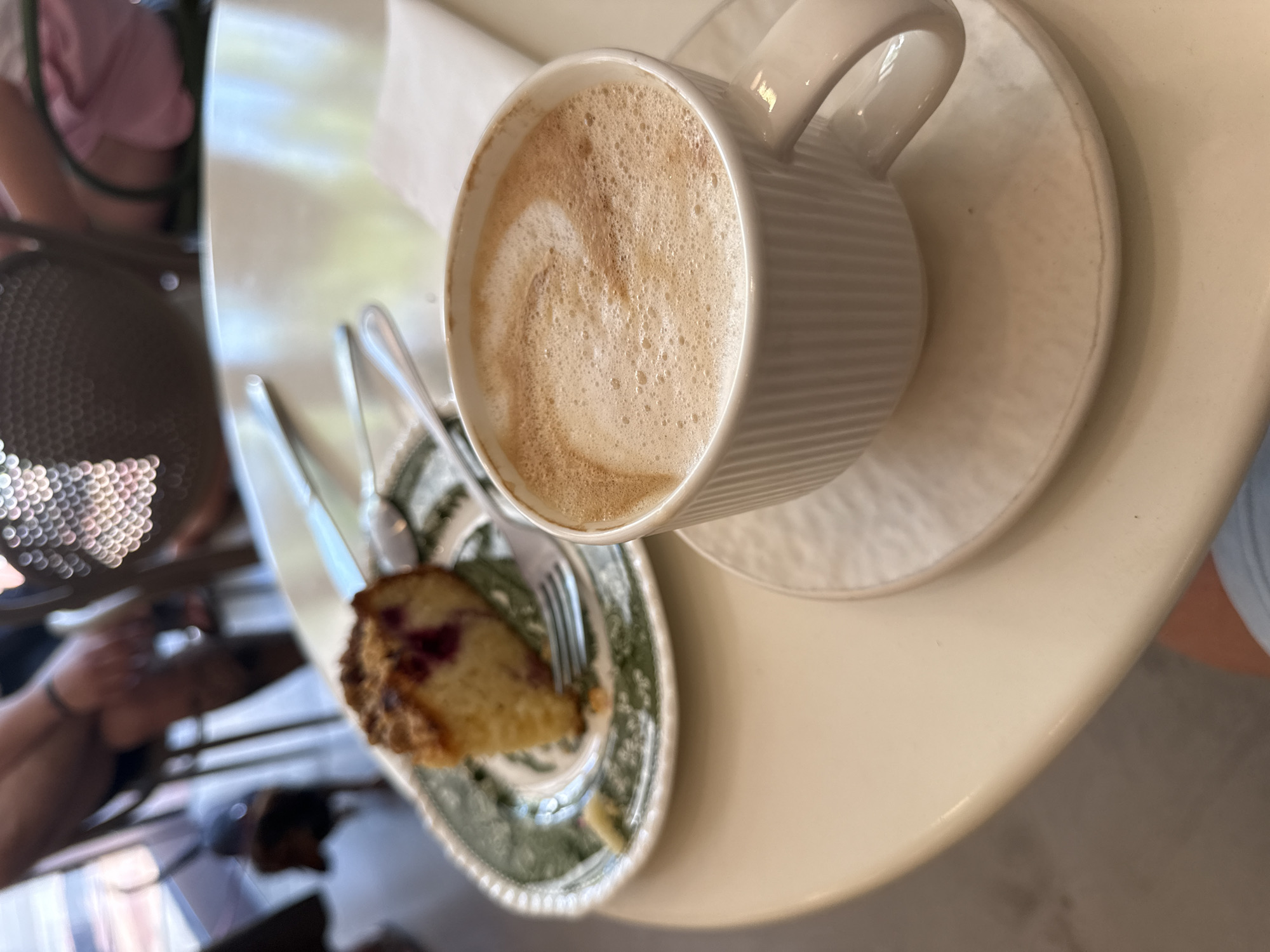 The image shows a close-up of a slice of cake and a cup of coffee on a table. The cake is on a plate with a green and white pattern. The coffee is in a white cup with a ribbed texture and a saucer. The table is light-colored and appears to be made of laminate. In the background, blurry figures of people are visible, suggesting a cafe or restaurant setting. The overall lighting is bright, creating a natural and casual atmosphere. The image does not provide any specific information about Budva, Montenegro, nor does it depict any actions or interactions that could be considered forensic in nature.
