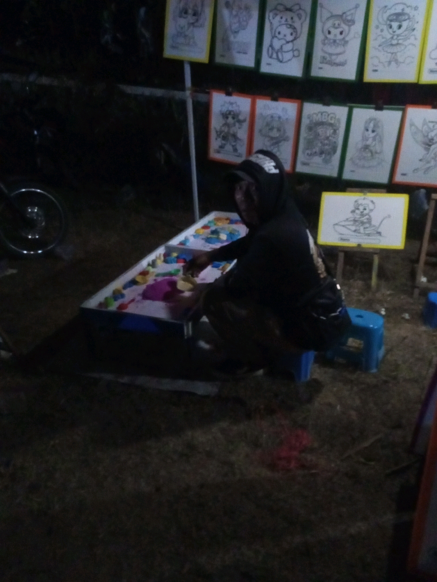A person wearing a black hooded jacket and a black baseball cap is kneeling beside a long, white rectangular table filled with colorful sand and sand molds. The person's hands are engaged in playing with the sand. Behind the table, a display rack holds several framed drawings of cartoon characters. To the left of the table, a portion of a motorcycle wheel is visible. The scene appears to be outdoors, possibly at a market or fair, given the display of drawings and the sand play activity. The ground is covered in grass and dirt. The overall lighting is dim, suggesting it is either dusk or nighttime. The location is identified as Ngadikerso, Indonesia.