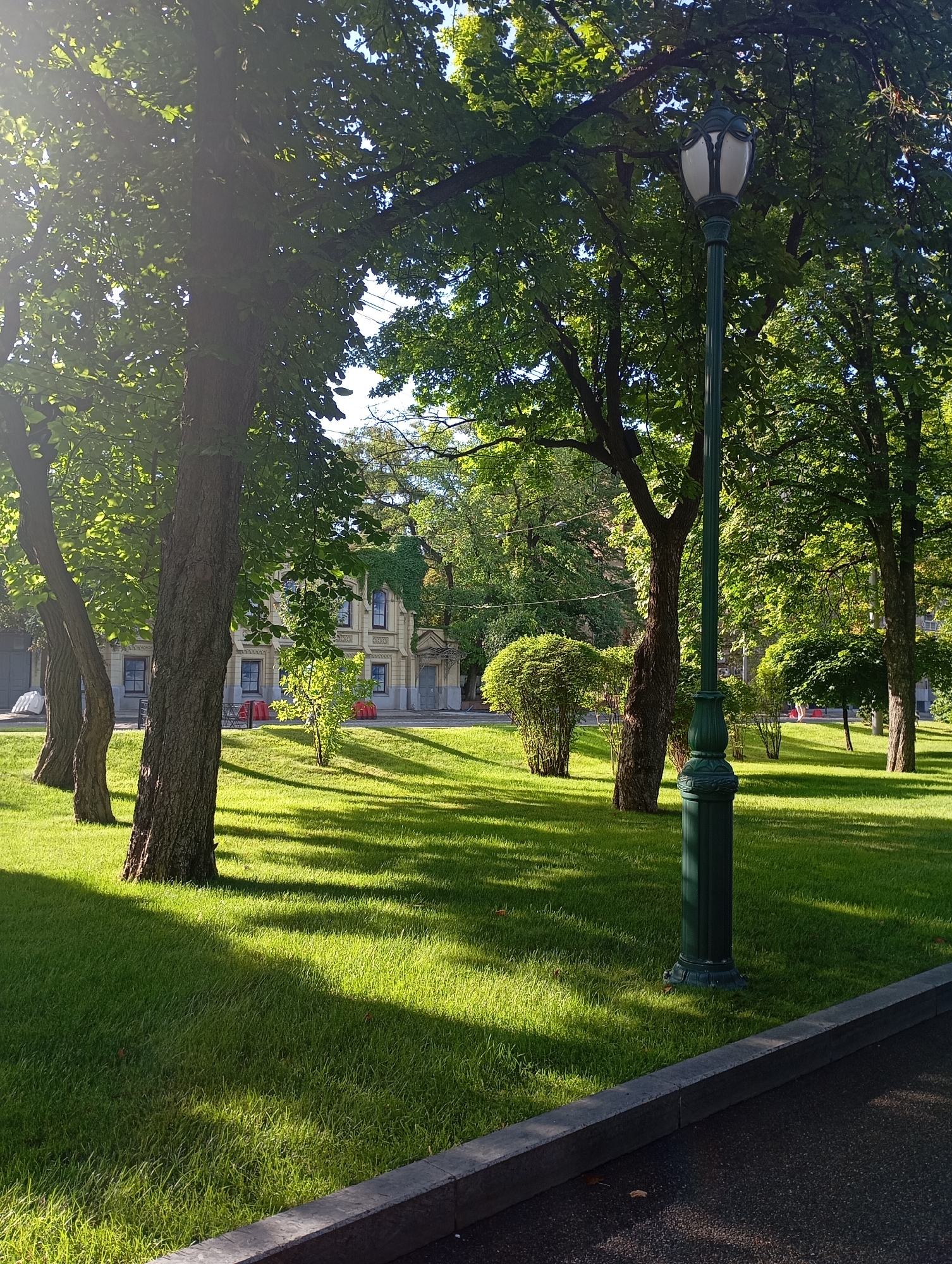 The image depicts a park in Kharkiv, Ukraine, featuring a lush green lawn, mature trees casting dappled shadows, and a classic lamppost in the foreground. In the background, a light-colored, classical-style building with multiple windows is visible. The overall scene is serene and bathed in sunlight, suggesting a pleasant day in an urban green space.