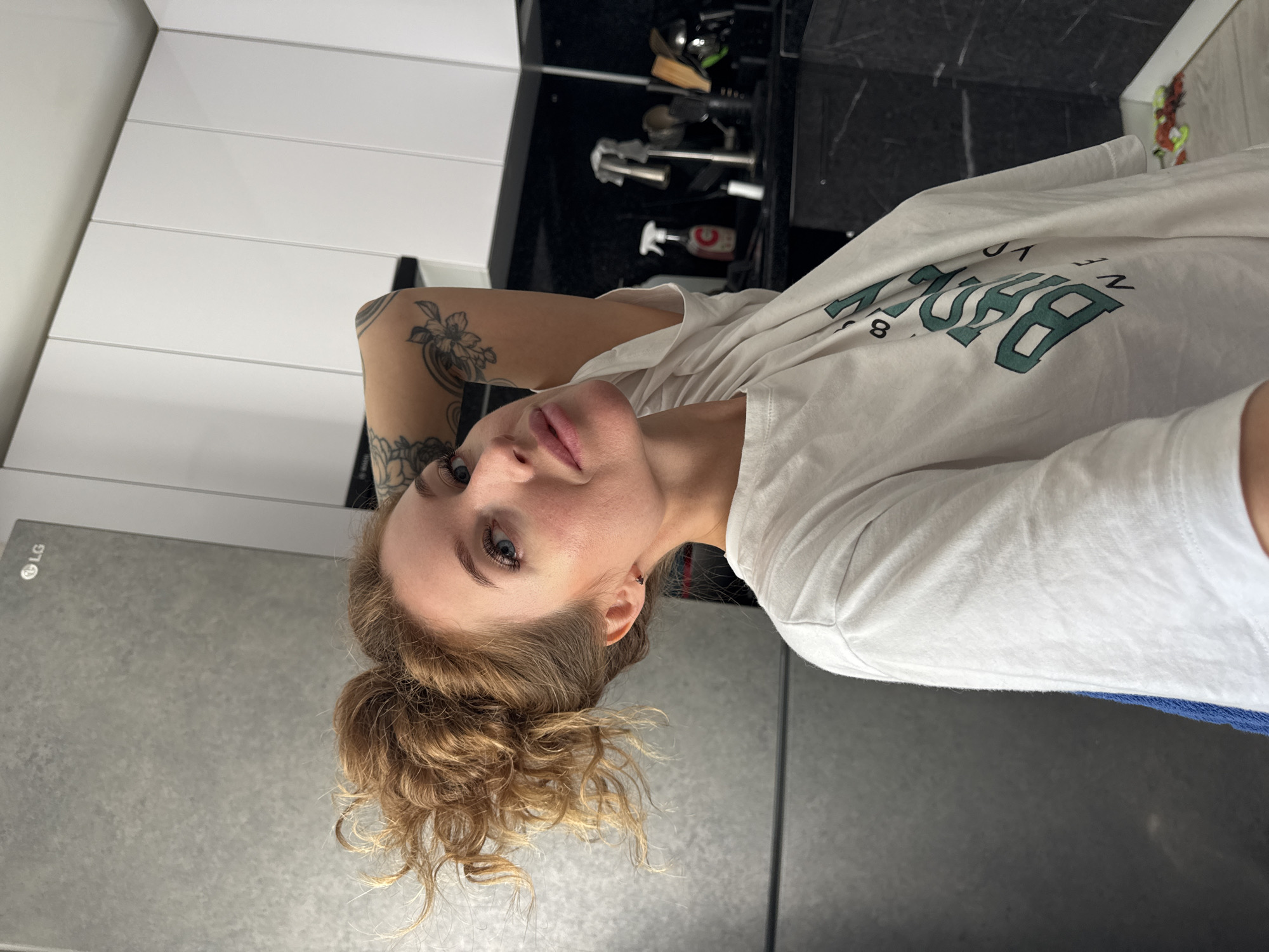 A woman with blonde curly hair pulled up in a messy bun is shown in a selfie taken in a kitchen. She is wearing a white t-shirt with green lettering that reads "BROOKLYN". The background shows a grey refrigerator on the left and white cabinets on the right. The kitchen also features black countertops and a faucet. The woman has tattoos on her arm and is looking at the camera with her hand behind her head. The location is the city of Chaikovsky, Russia.