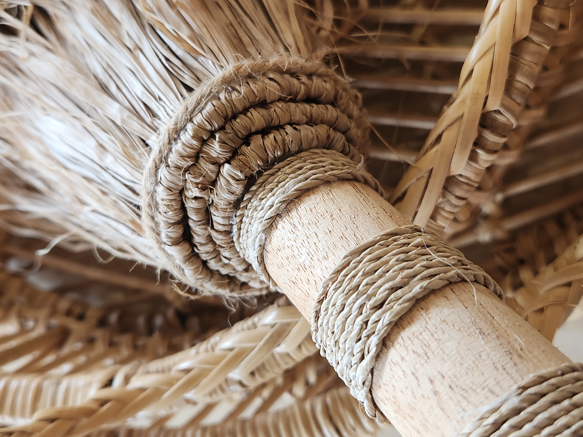 The media file is a close-up, shallow-depth-of-field photograph focusing on the textured details of a light-colored, wooden cylindrical object wrapped with natural fiber ropes and twine. The object appears to be part of a larger structure, possibly an umbrella or a piece of furniture, as suggested by the blurred background which shows woven, straw-like material.
The foreground clearly shows the wooden cylinder with a visible wood grain texture. Two distinct rope wrappings are present. The upper wrapping is thicker and more densely woven, creating a layered, almost braided effect around the cylinder. The lower wrapping consists of multiple strands of thinner rope, twisted together and wound around the cylinder in a spiral pattern. The color of the ropes is a natural, light tan or beige.
In the upper left portion of the frame, a bundle of straw or dried grass-like material fans out from the wooden cylinder, secured by the upper rope wrapping. This material is pale and appears somewhat frayed at the edges.
The background is out of focus but reveals a repeating pattern of thin, woven strips, likely also made from natural fibers, suggesting an architectural or decorative element. The lighting appears to be natural, casting soft shadows and highlighting the textures of the materials. There is no text visible in the image. The overall impression is one of natural materials and handcrafted construction, evoking a sense of rustic or tropical decor. The location context is Maitland, United States of America.