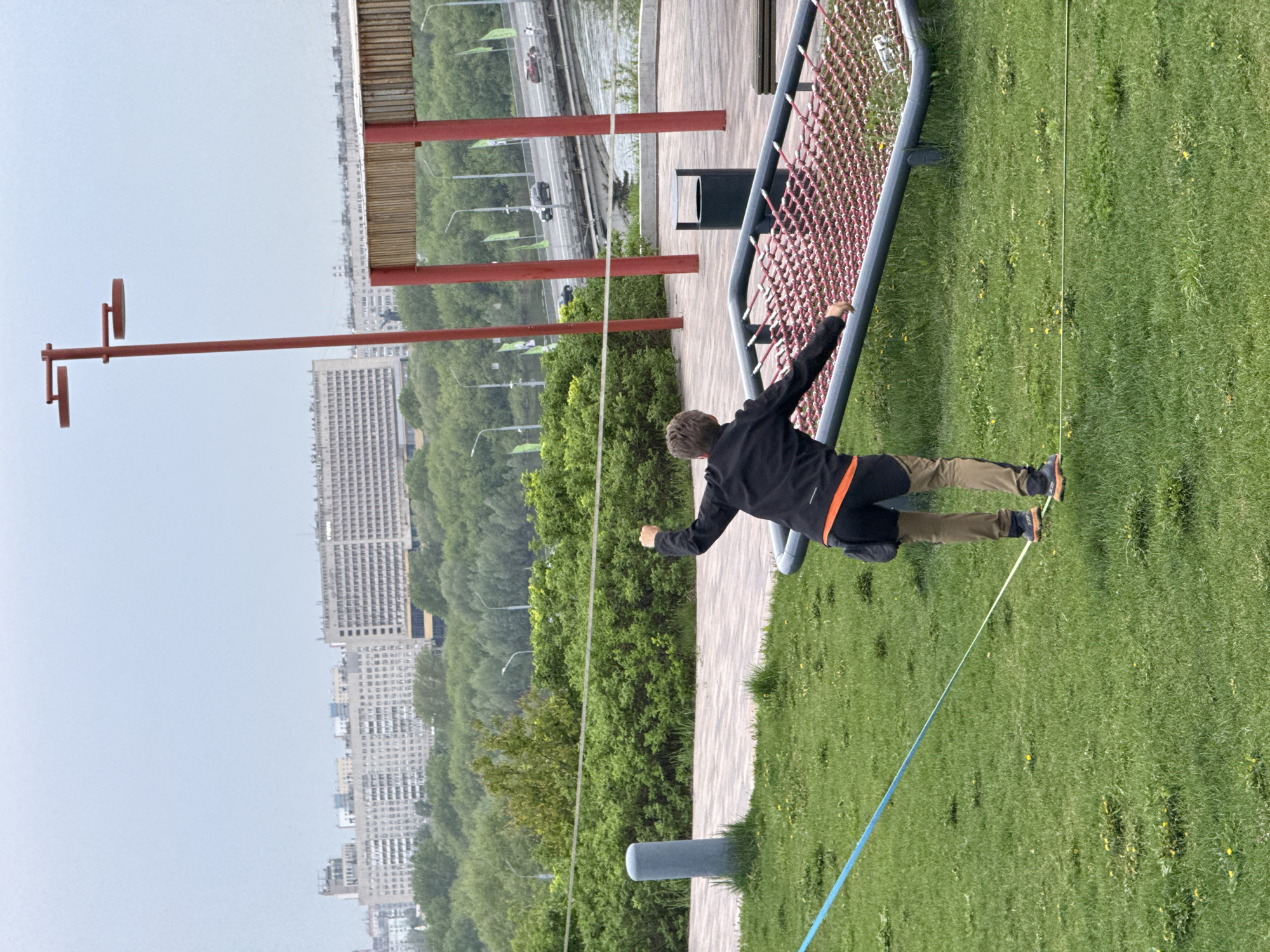 A high-angle, outdoor view of a person tightrope walking in what appears to be a park in Nizhny Novgorod, Russia. The person, dressed in a dark jacket with orange accents and tan pants, is positioned on a white tightrope strung across a grassy area. Their arms are extended for balance. A blue slackline is visible beneath the white tightrope. The park setting includes trees, a grey playground structure featuring a red net, a grey trash receptacle, and a paved walkway. In the distance, buildings and trees constitute the cityscape of Nizhny Novgorod. A red-pillared overpass or structure is also visible. The sky is overcast. The photograph is rotated counter-clockwise.