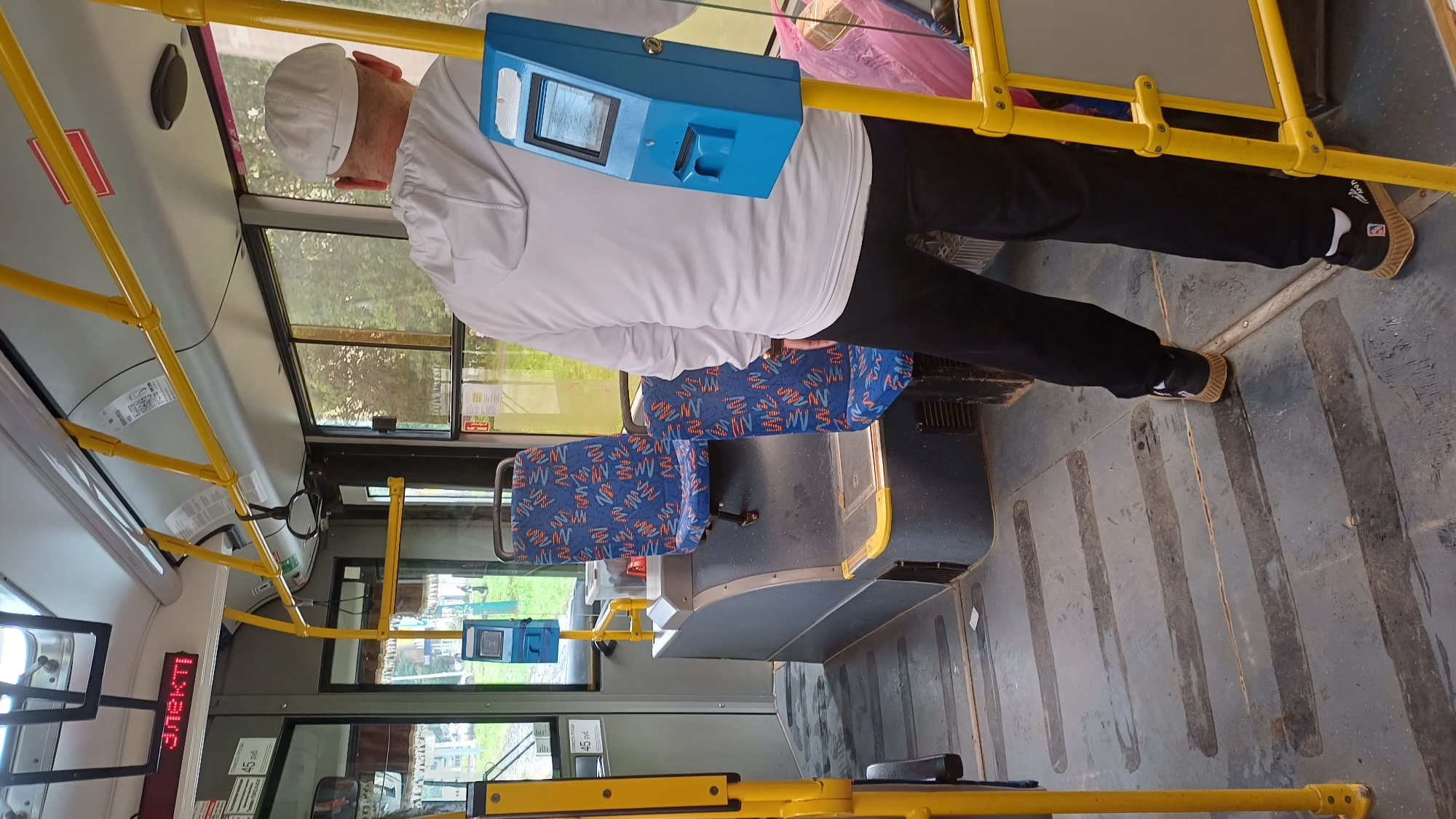 The image depicts the interior of a public bus in Izhevsk, Russia. A man wearing a white hooded sweatshirt, black pants, and a white cap stands near a blue ticket vending machine. The bus features blue seats with a patterned design, yellow handrails, and a grey floor. A digital display above the seats shows "ЭЛЕКТР" in red letters, likely indicating "Electric." Another sign shows "45 руб.", denoting a fare of 45 rubles. The environment suggests an urban transit setting.