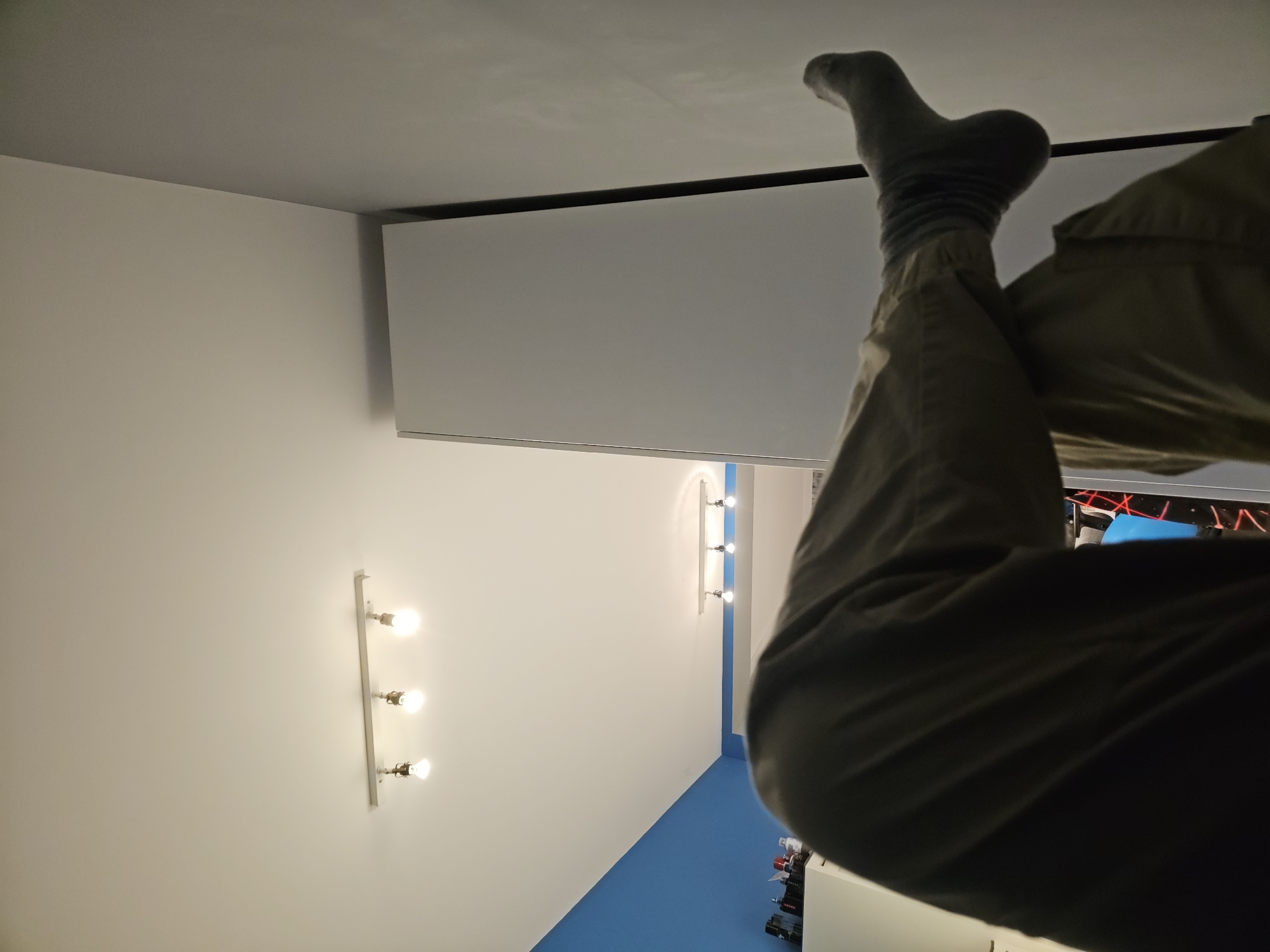 The image shows a room with white walls and a blue accent wall. A track light fixture with three bulbs is visible on the white wall. An individual's legs, clad in khaki pants and a gray sock, are positioned towards the upper right of the frame, obscuring a television or computer monitor in the background. The overall perspective is from a low angle, looking upwards towards the ceiling. The location in Sofia, Bulgaria cannot be determined from the image.