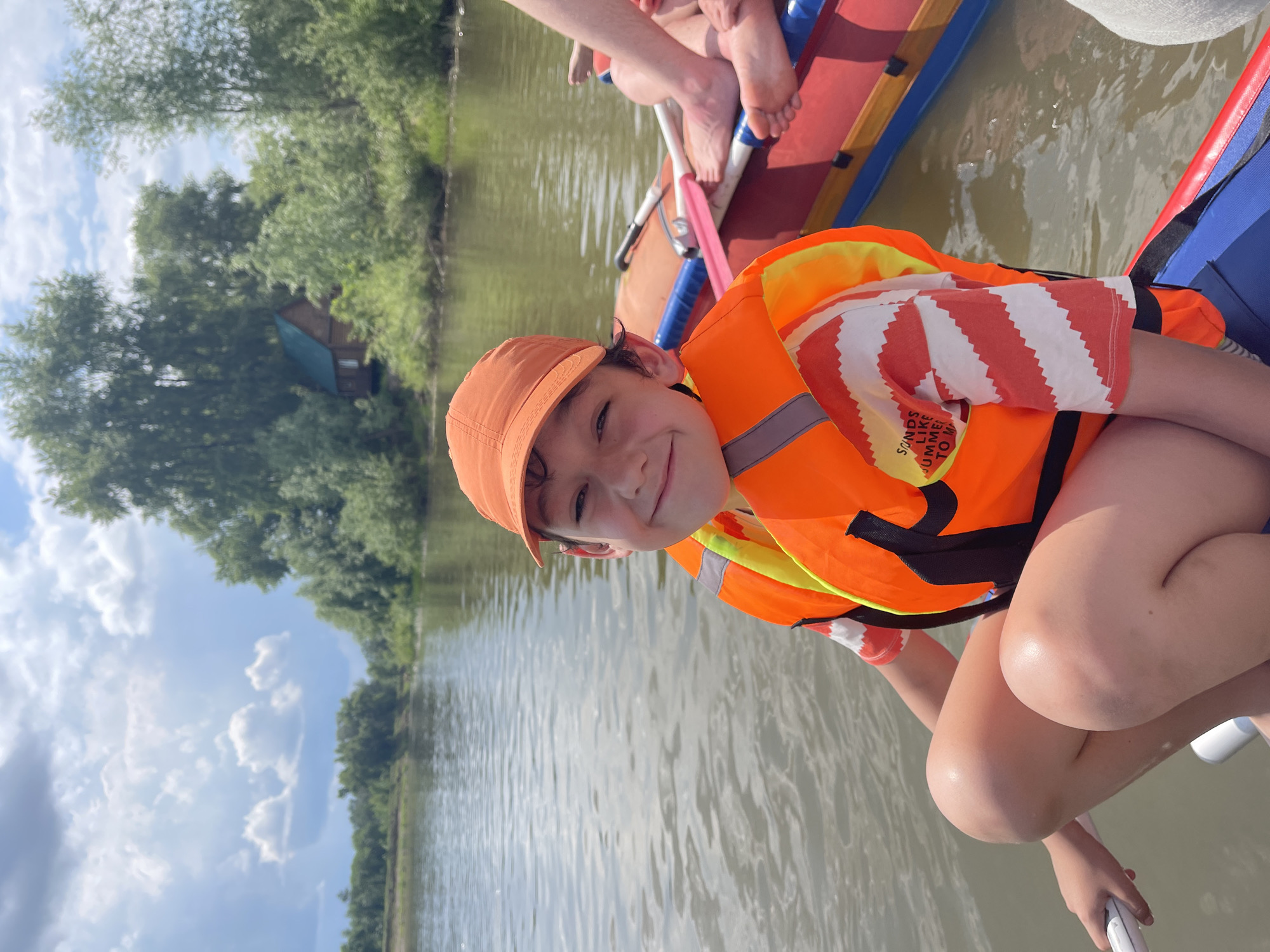 A young boy wearing an orange baseball cap and an orange life vest smiles at the camera while kneeling on a raft. He has on a white and orange striped shirt. The raft is on a body of water surrounded by lush green trees. In the background, a small wooden house with a blue roof can be seen among the trees. The sky is blue with fluffy white clouds. The image appears to be taken on a sunny day. The raft has other people on it, whose legs and feet are visible. The setting is likely a natural environment, possibly a river or lake, in Нежинка, Russia. The boy's expression suggests happiness and excitement. The life vest has reflective stripes. Partially visible text on the boy's shirt reads "SOUNDS LIKE SUMMER TO M".
