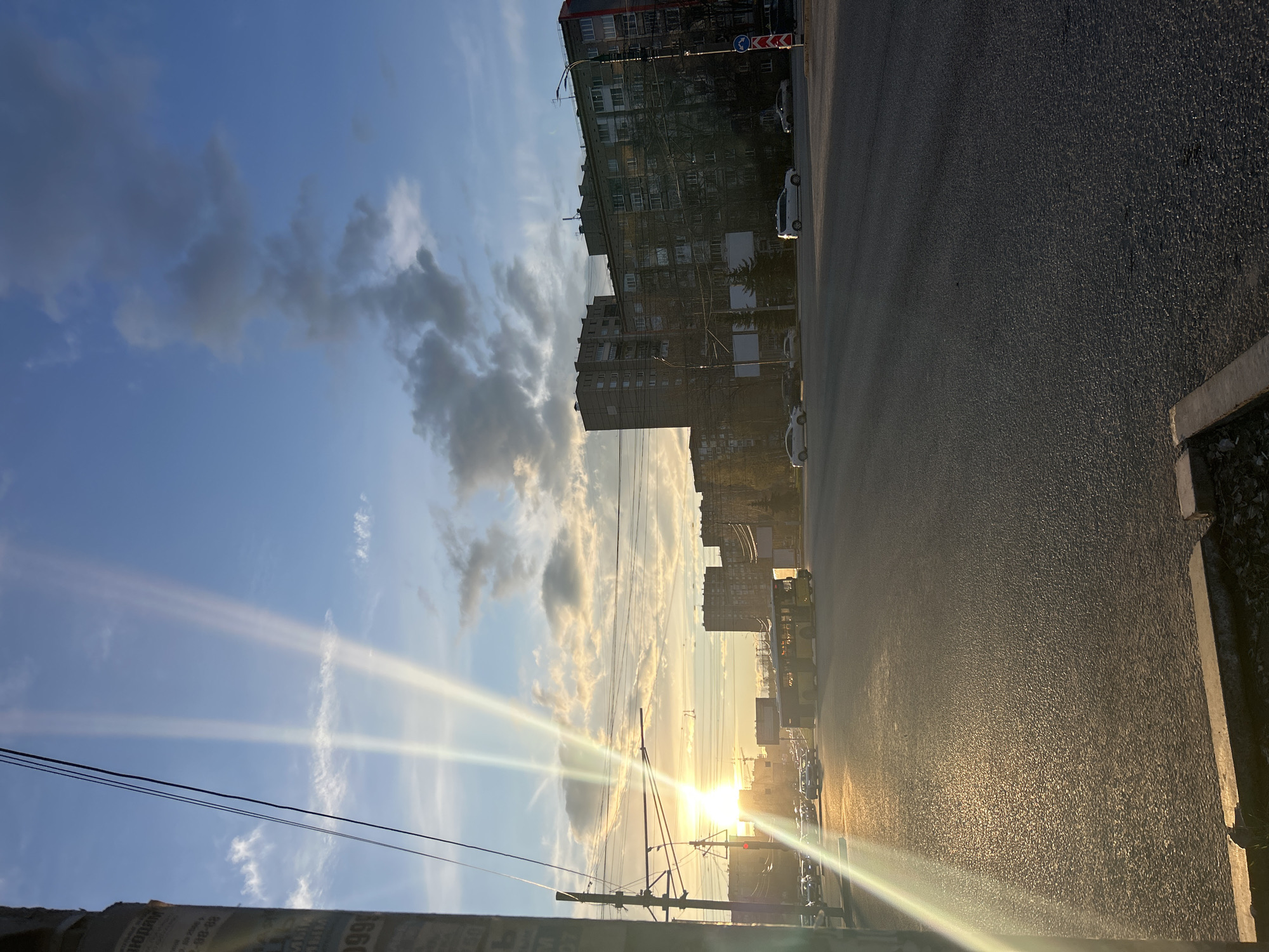 This image depicts an urban street scene in Izhevsk, Russia, captured during a vivid sunset. The low sun, positioned towards the left of the frame, casts brilliant rays that pierce through the sky, creating a strong lens flare and a warm, golden glow across the landscape. The sky is a mix of clear blue and scattered clouds, some illuminated by the setting sun.
On the left side of the street, several multi-story buildings, consistent with residential or commercial architecture, rise into the sky. One building features scaffolding, suggesting ongoing construction or renovation. Numerous utility poles and overhead wires crisscross the mid-ground, a common feature of urban infrastructure. Traffic signs, including a red and white chevron and a blue circular sign, are visible on a pole near one of the buildings.
The wide asphalt street occupies the right and foreground of the image, reflecting the golden light of the sunset, which creates a textured shimmer on its surface. Several cars, including distinct white vehicles, are parked along the street. In the distance, other vehicles and what appears to be a red traffic light are discernible.
In the immediate foreground on the left, a post or pillar is partially visible, covered with torn paper and fragments of text. Recognizable elements include "Билайн" (Beeline, a telecommunications company), "Ремонт" (Repair), and partial phone numbers like "30 48-32" and "999". No people are visible in the scene, contributing to a tranquil, end-of-day atmosphere.